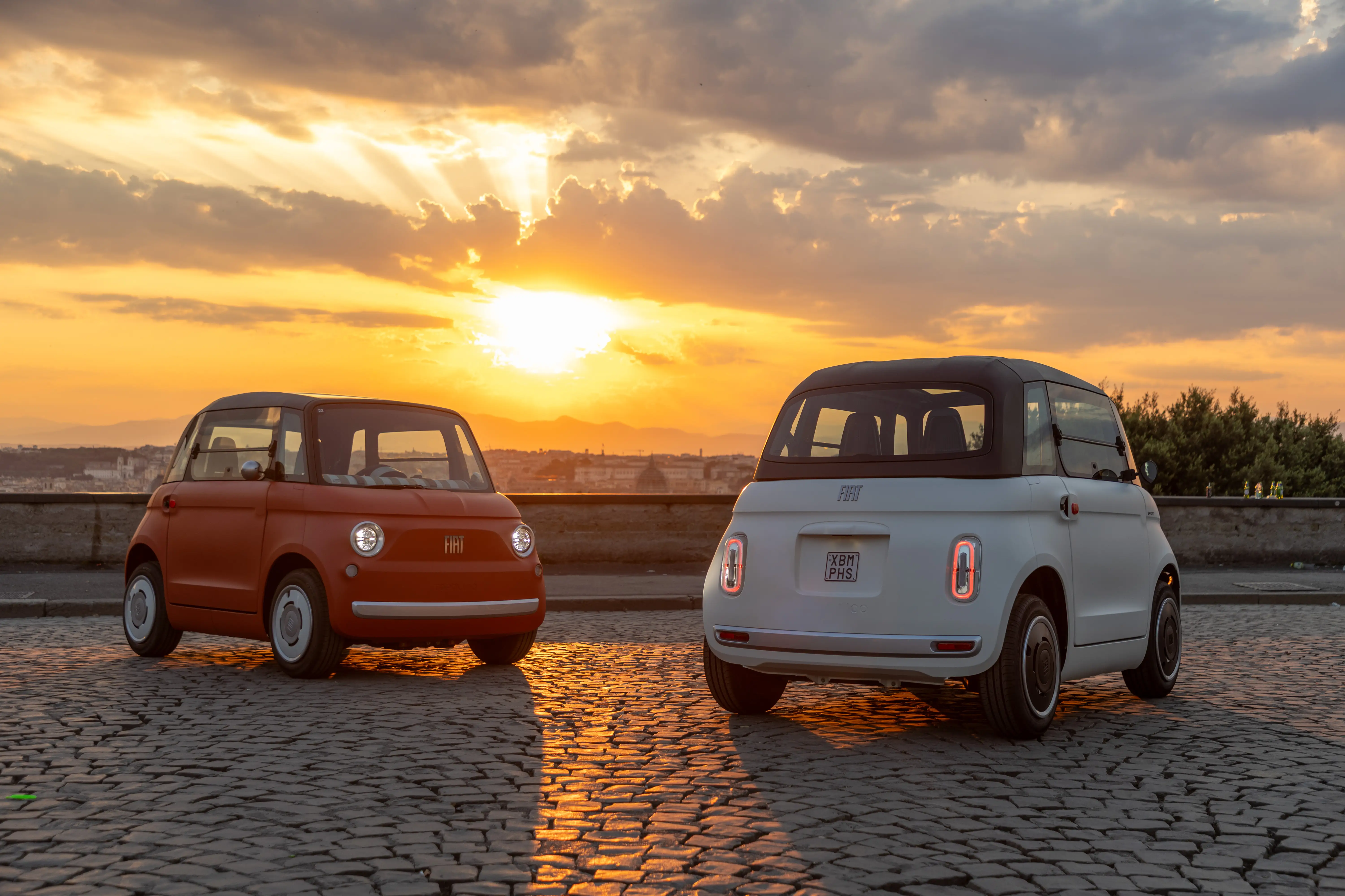 Two Fiat Topolinos are parked on a brick road during a sunset.
