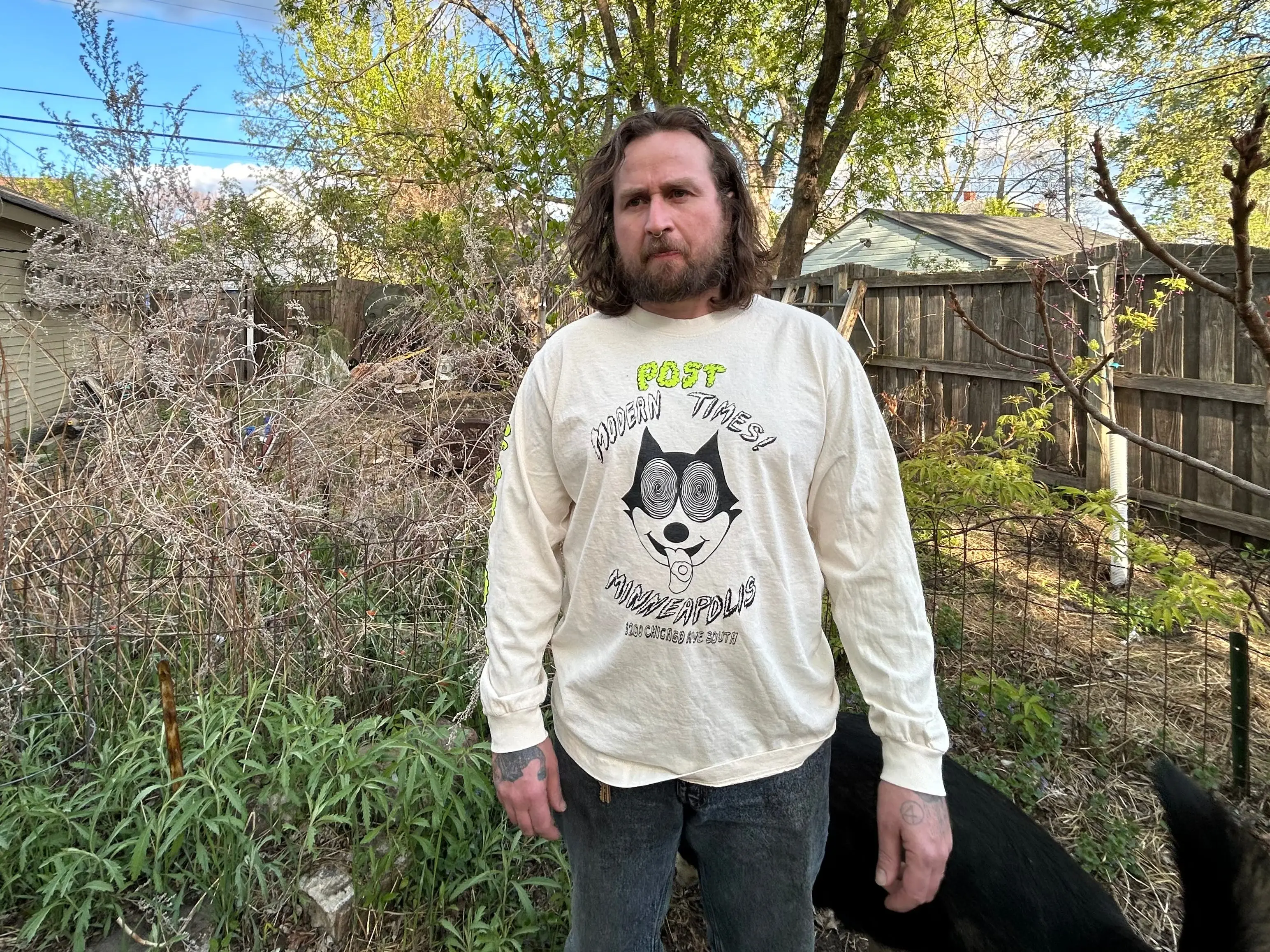 Dylan Alverson, the owner of the Post Modern Times stands in front of a tree wearing merch from his restaurant.