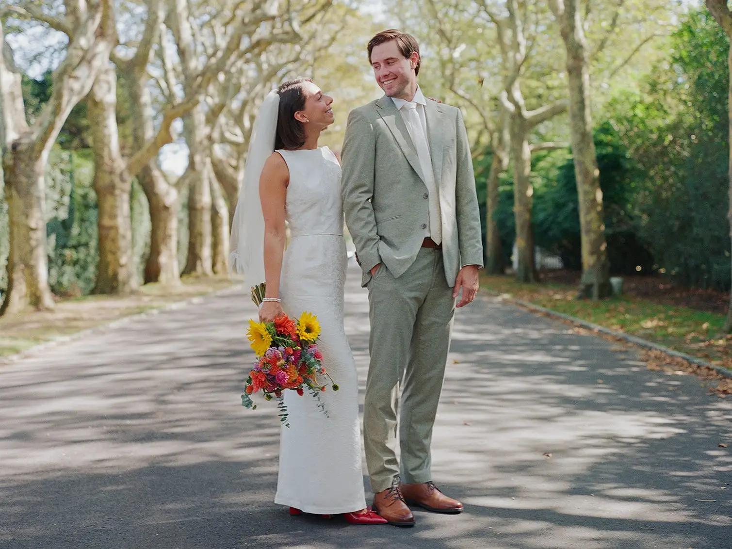 A bride and groom smile at each other on a tree-lined street.
