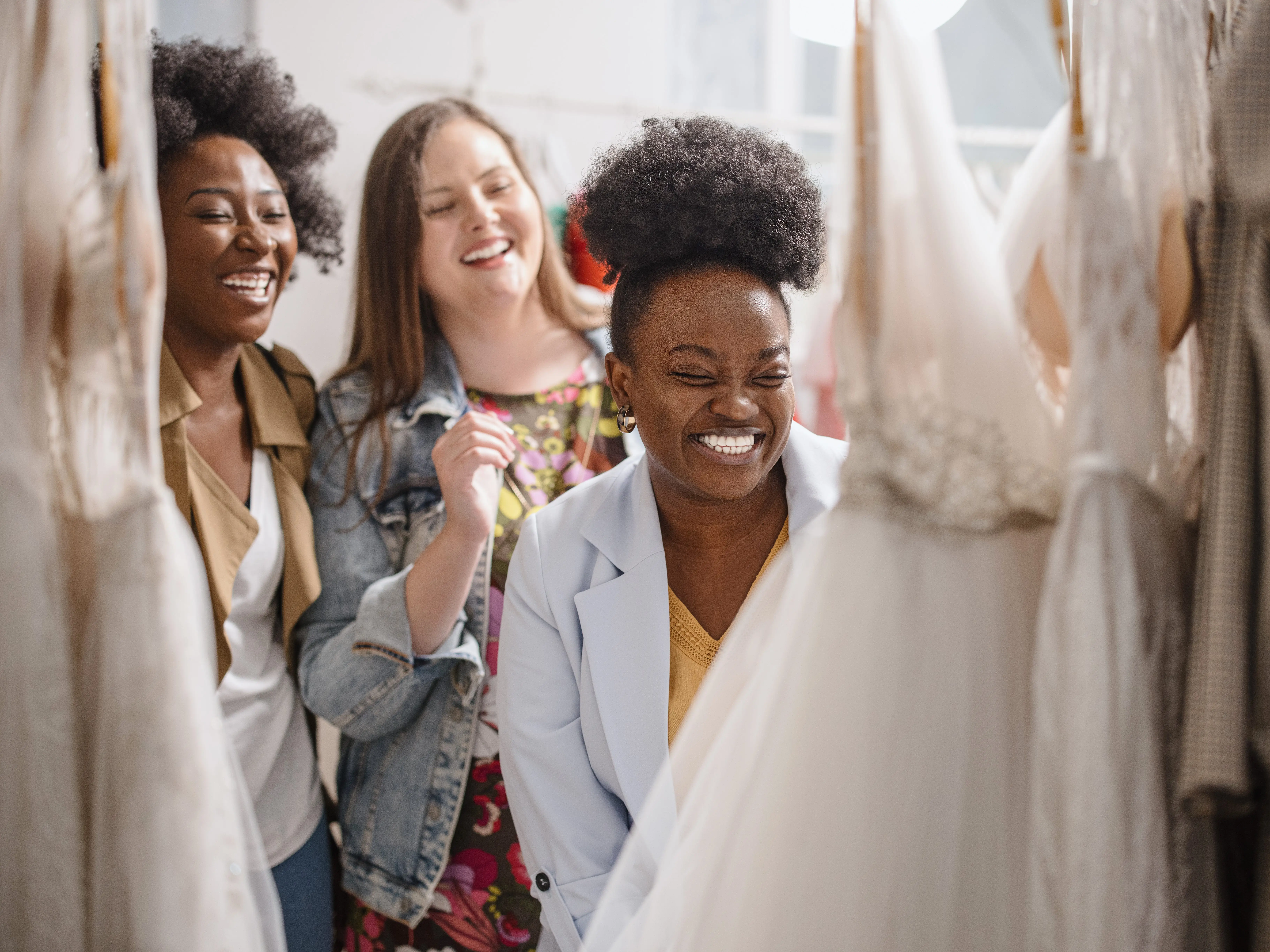 Three women smiling while looking at a wedding dress on a rack.