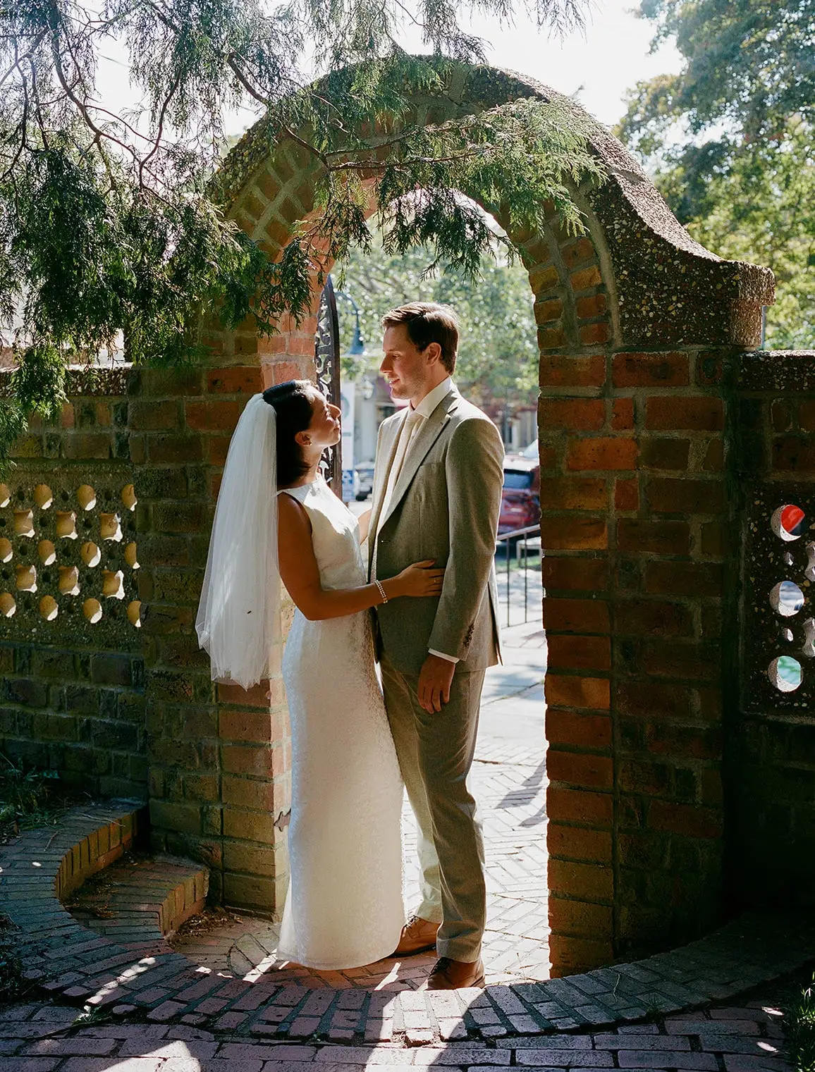 A bride and groom embrace and smile at each other under a brick archway.