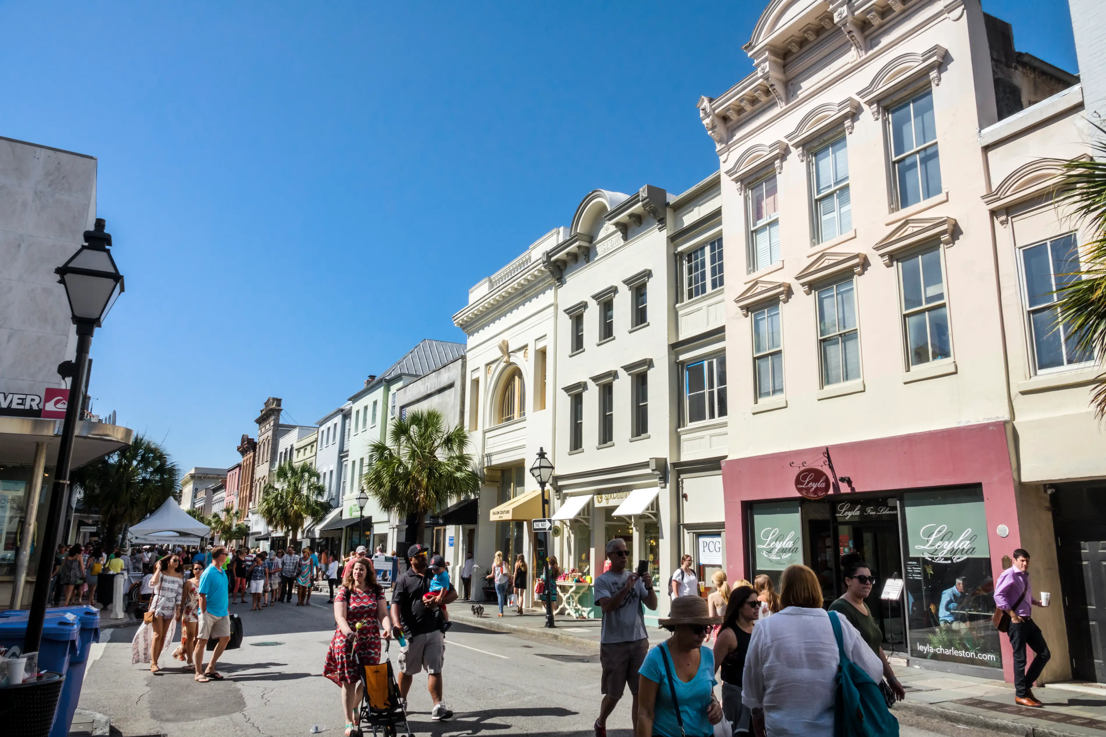 Pedestrians walk past pastel historic storefronts on a sunny street with palm trees and shop awnings.
