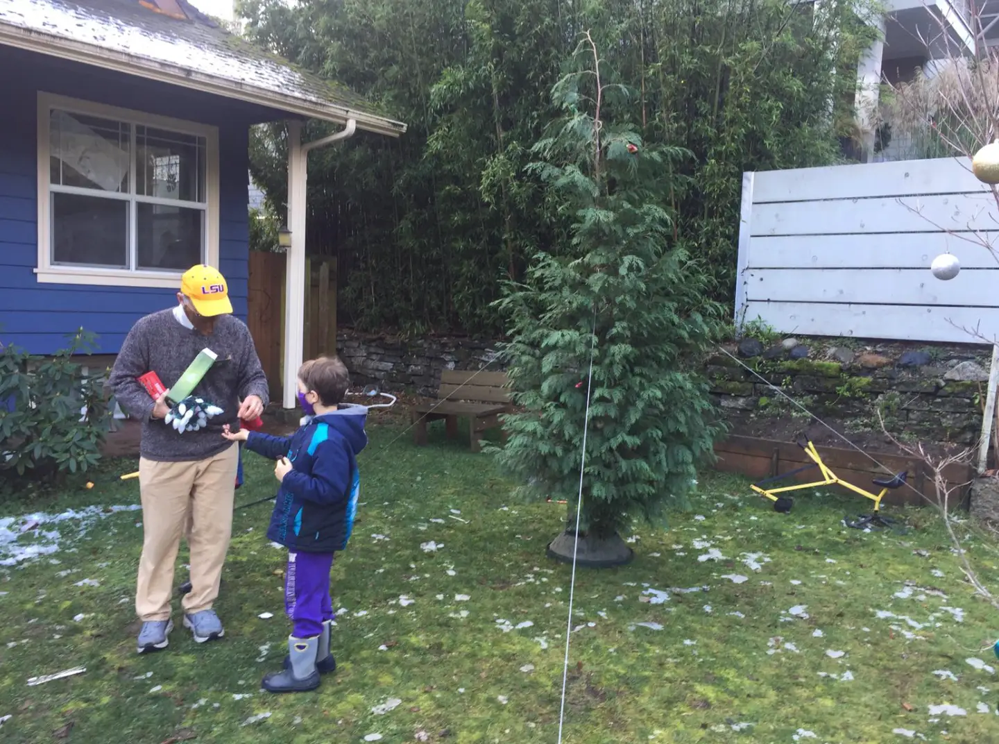 Two people stand in a backyard near a large decorated evergreen tree and a blue house.