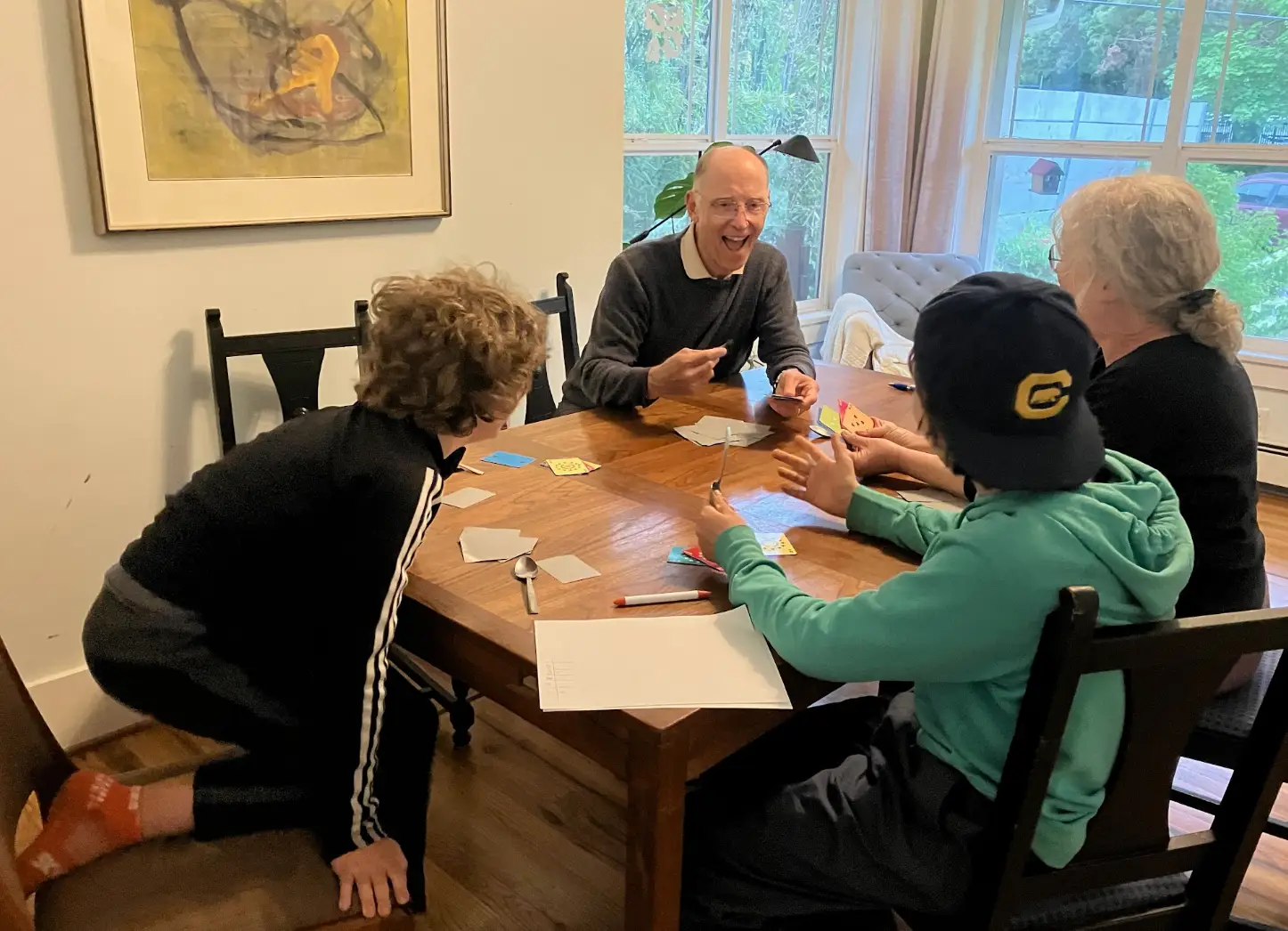 Four people sit around a wooden dining table with cards, paper, markers, and small craft materials near large windows.