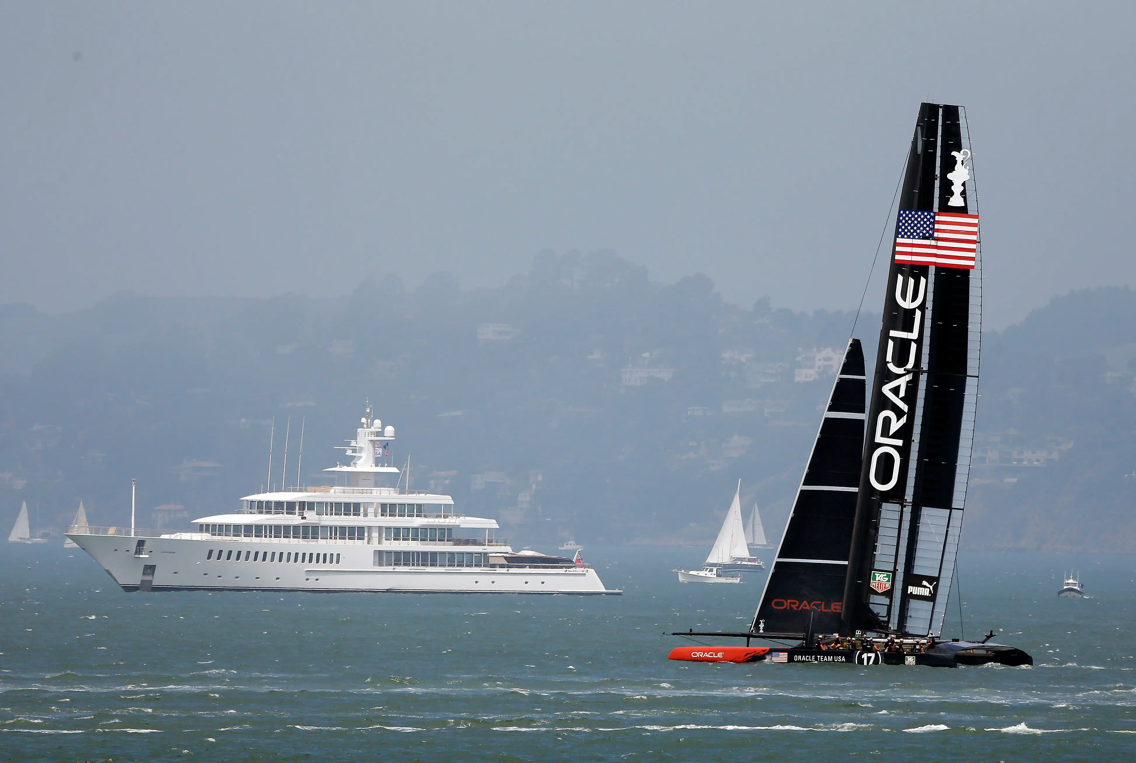 Oracle Team USA racing catamaran sails past a large white yacht and smaller boats on a hazy bay.