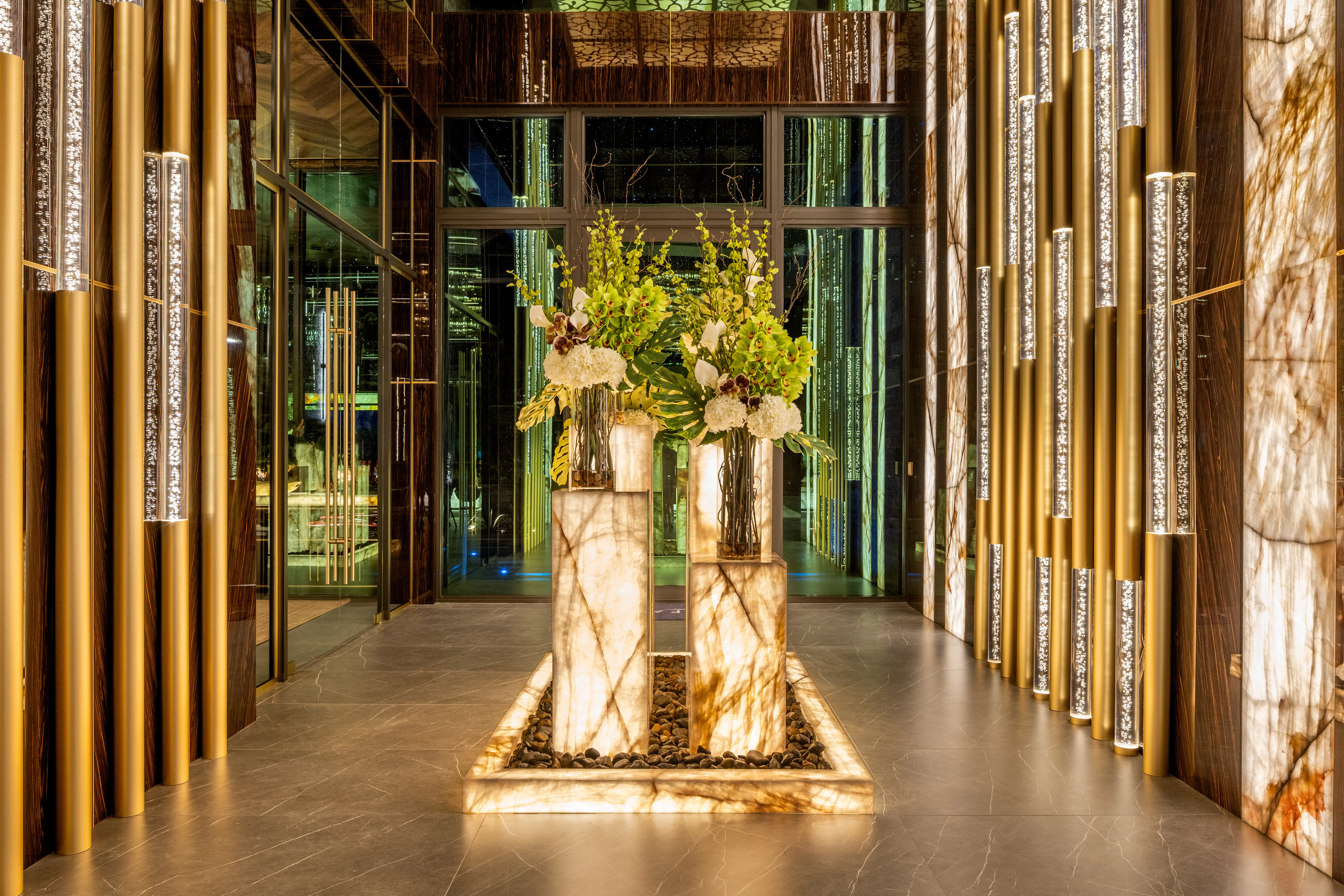 Luxurious illuminated lobby with marble columns, gold vertical light fixtures, glass doors, and large floral arrangements.