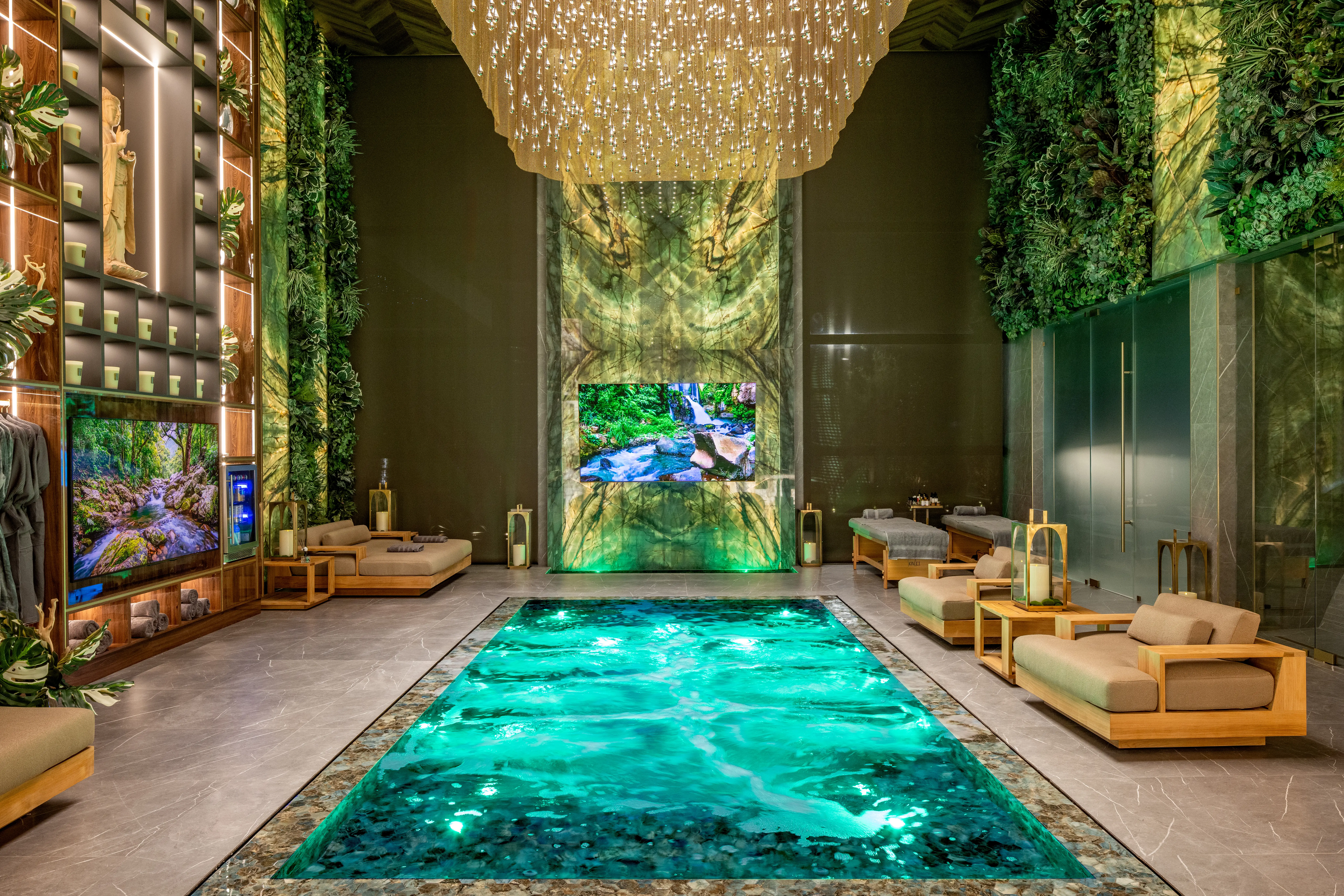 Luxurious indoor pool room with a large chandelier, green walls, lounge seating, and screens showing forest scenes.