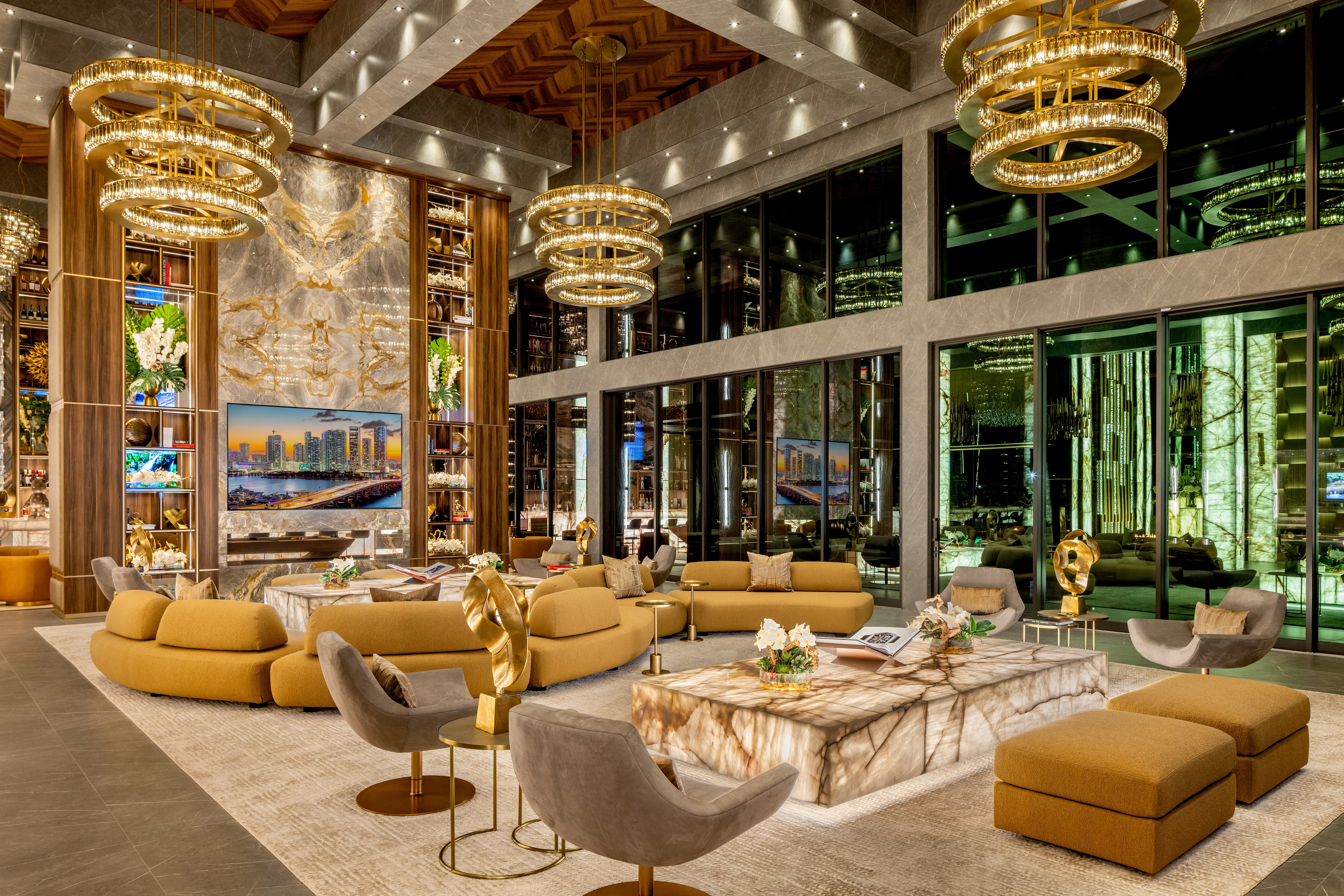 Luxurious high-ceiling lounge with gold chandeliers, marble tables, curved seating, large windows, and decorative shelving.