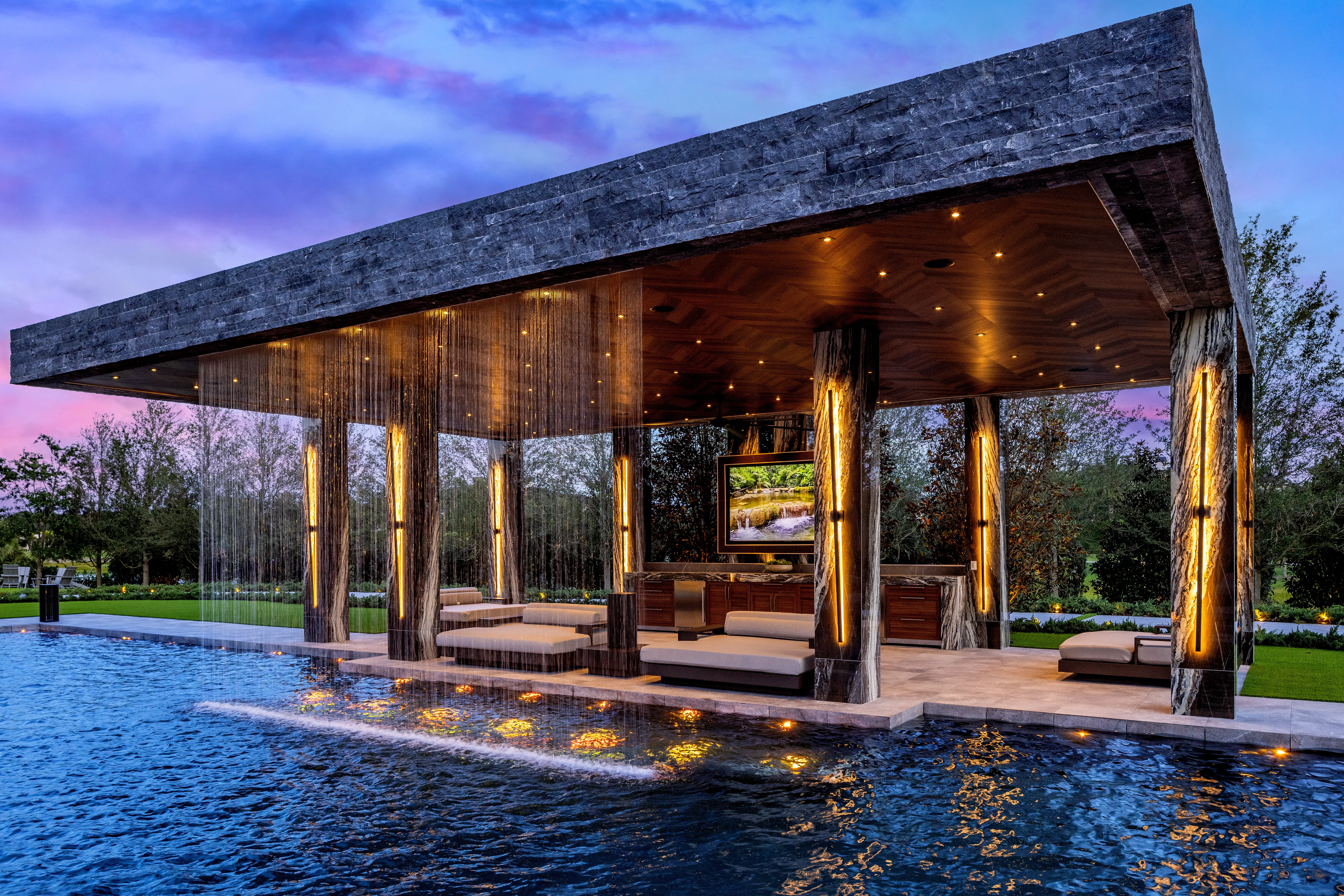 Modern illuminated poolside pavilion with water curtains, seating, and a television at twilight.
