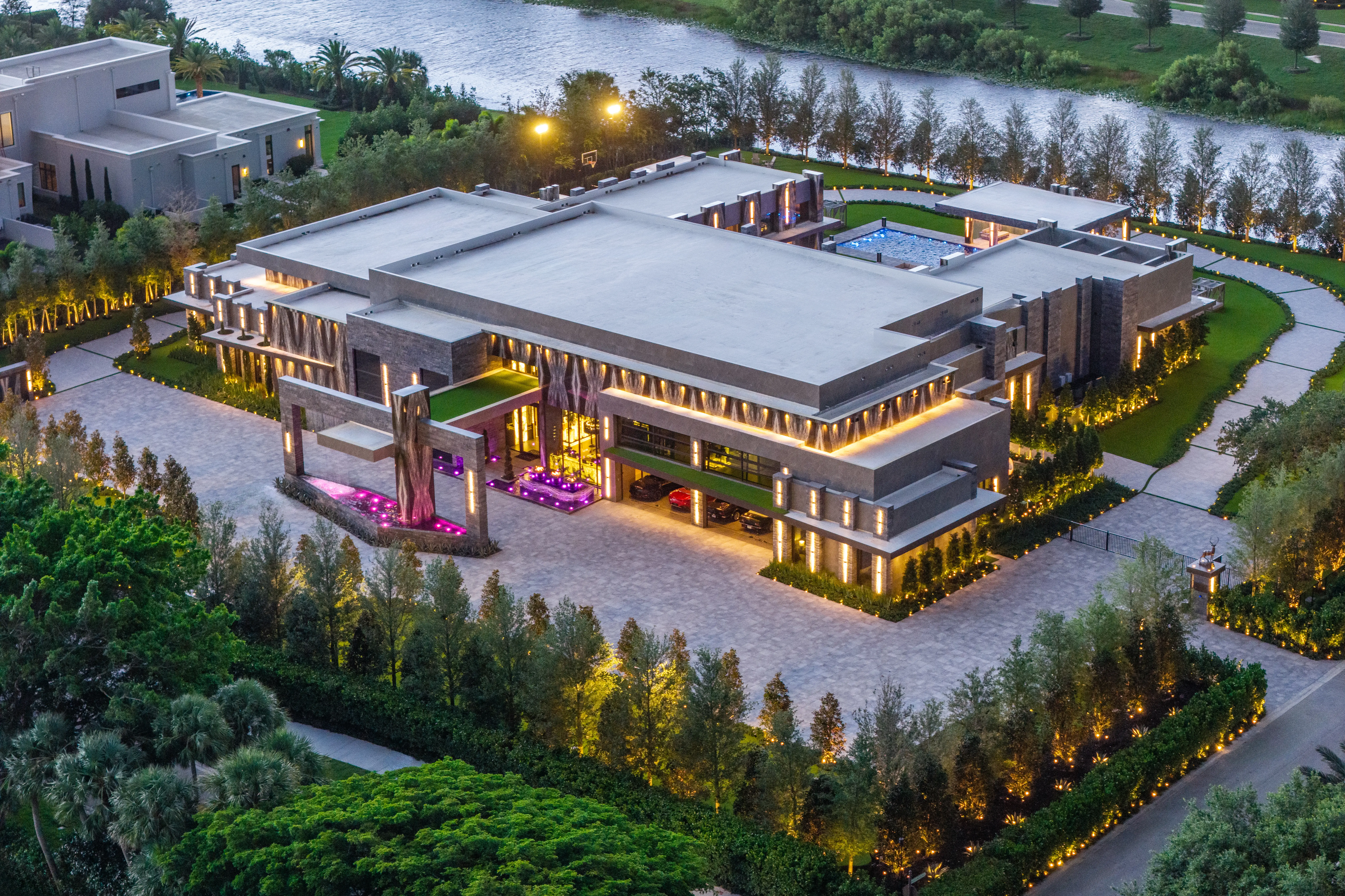 Aerial view of a large modern waterfront mansion illuminated at dusk with landscaped grounds and a pool.