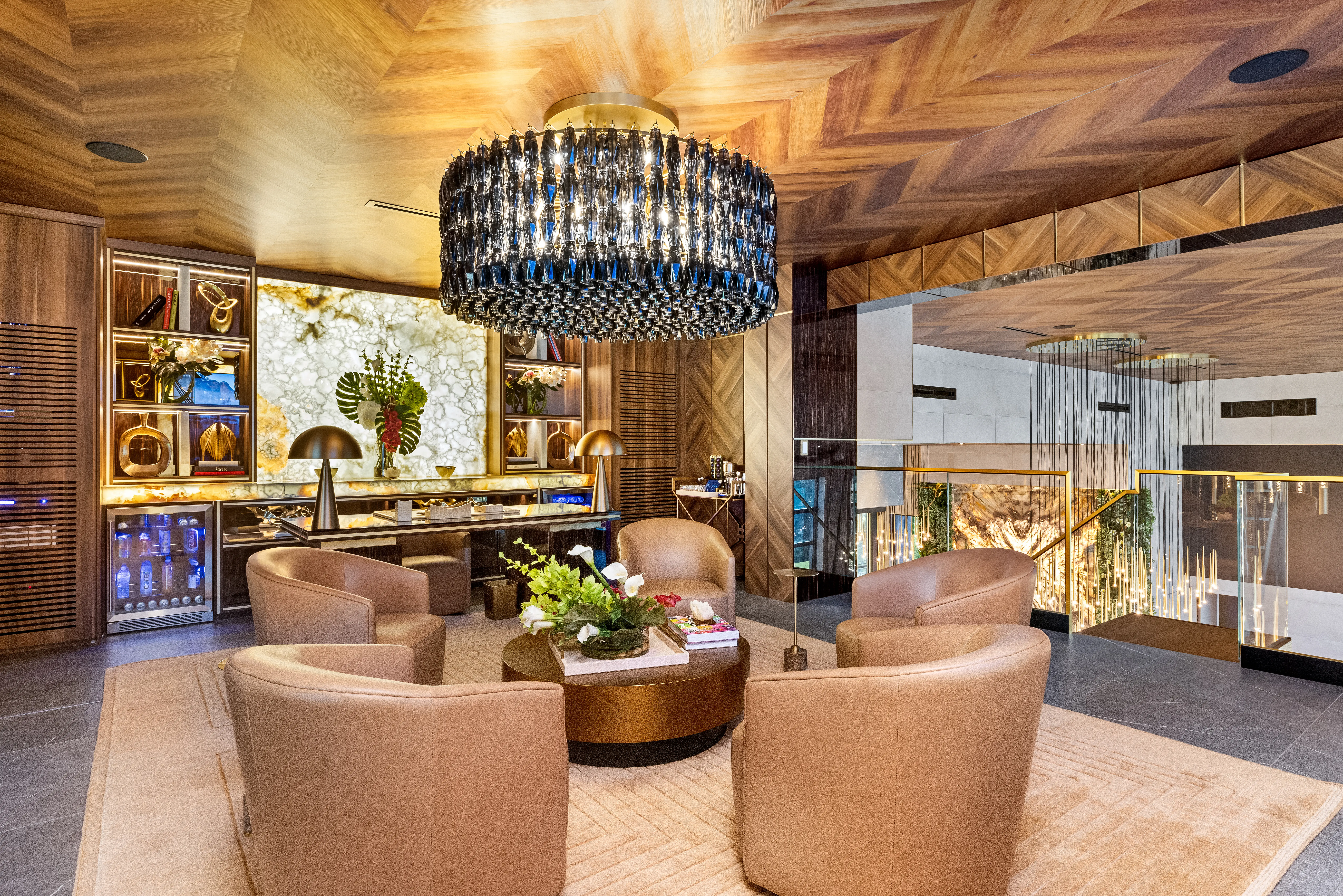 Luxury lounge interior with leather chairs, round table, illuminated shelving, wood ceiling, and large crystal chandelier.
