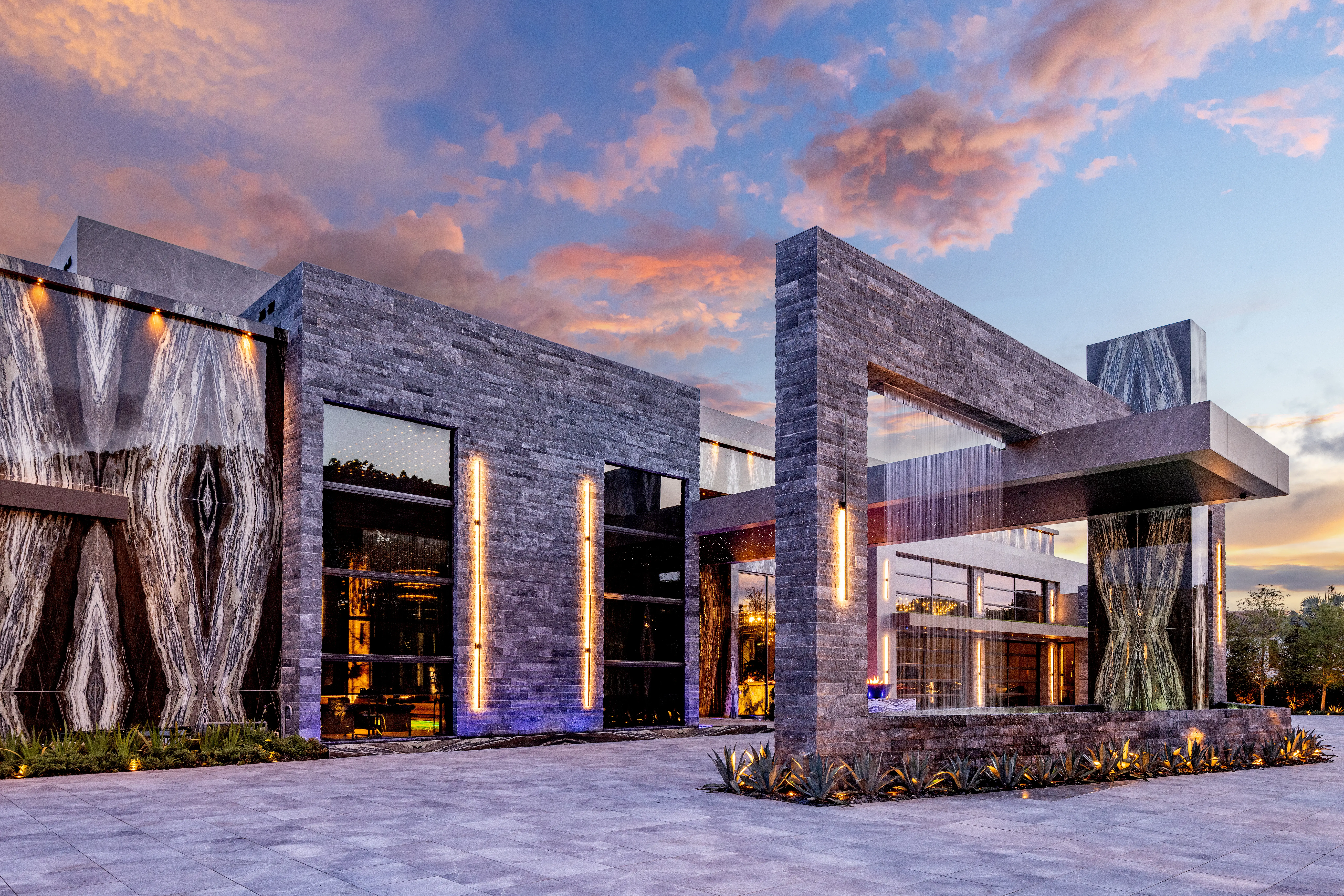 Modern stone-and-glass building exterior with illuminated vertical lights under a colorful sunset sky.