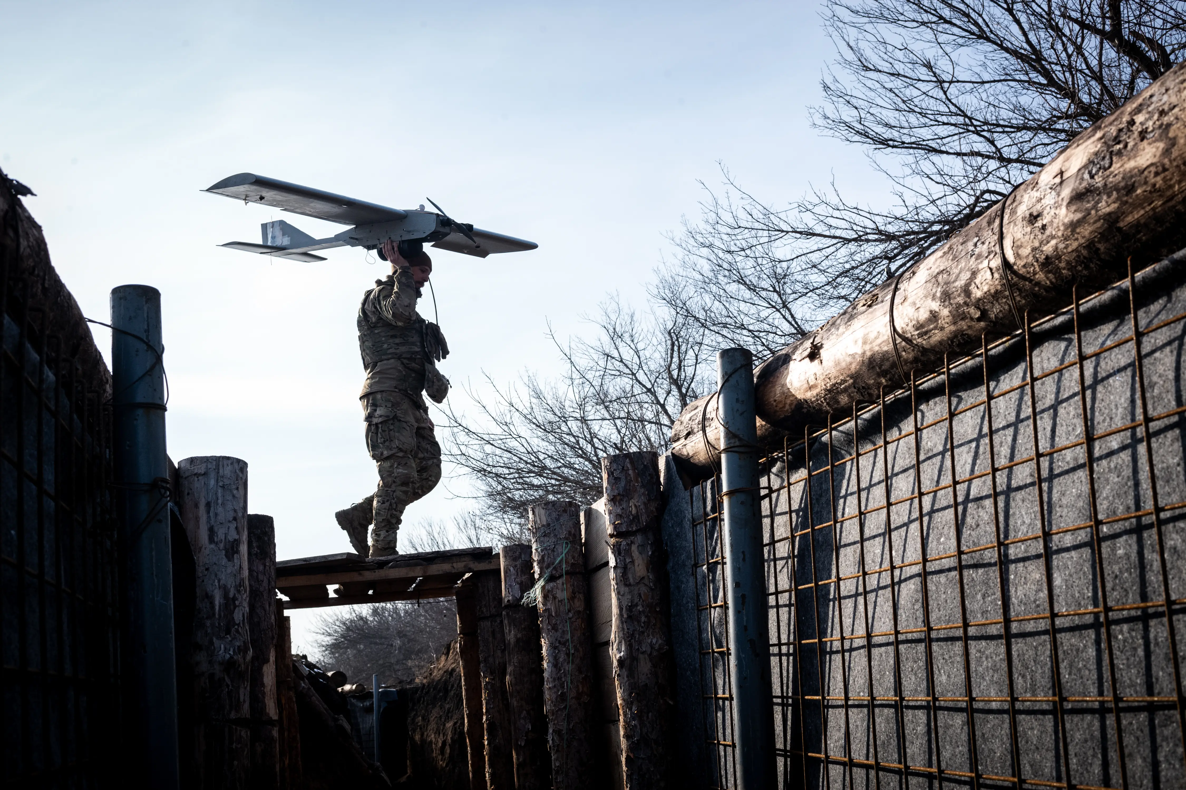 A figure walks across a bridge over a trench holding a drone in the air against a blue sky and bare trees
