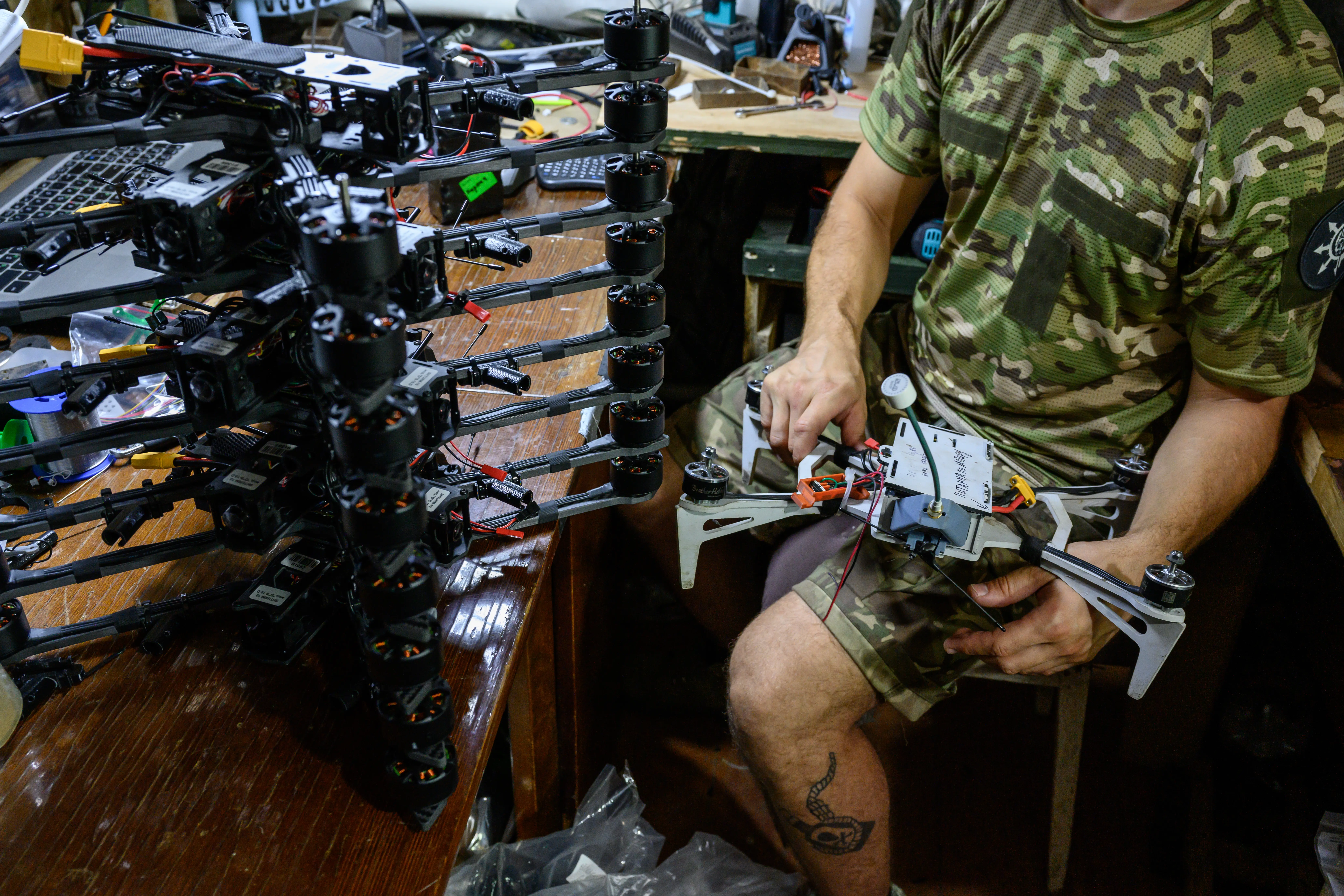 A man in camouflage gear and shorts sits on a seat holding a small drone with a stack of drones on a wooden surface beside him