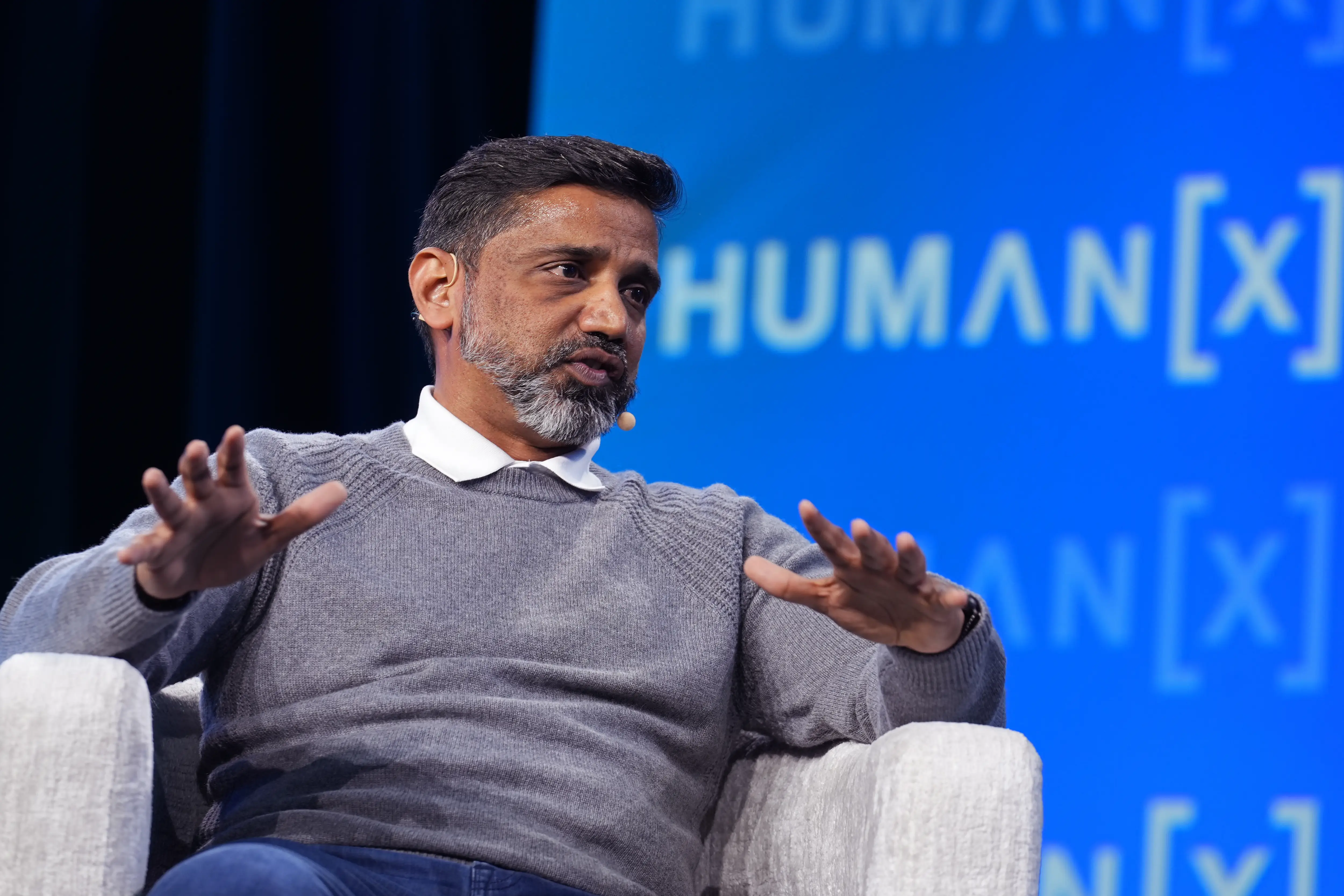 Uber CTO Praveen Neppalli Naga speaks on a stage while sitting in a white chair and wearing jeans and a grey sweater.