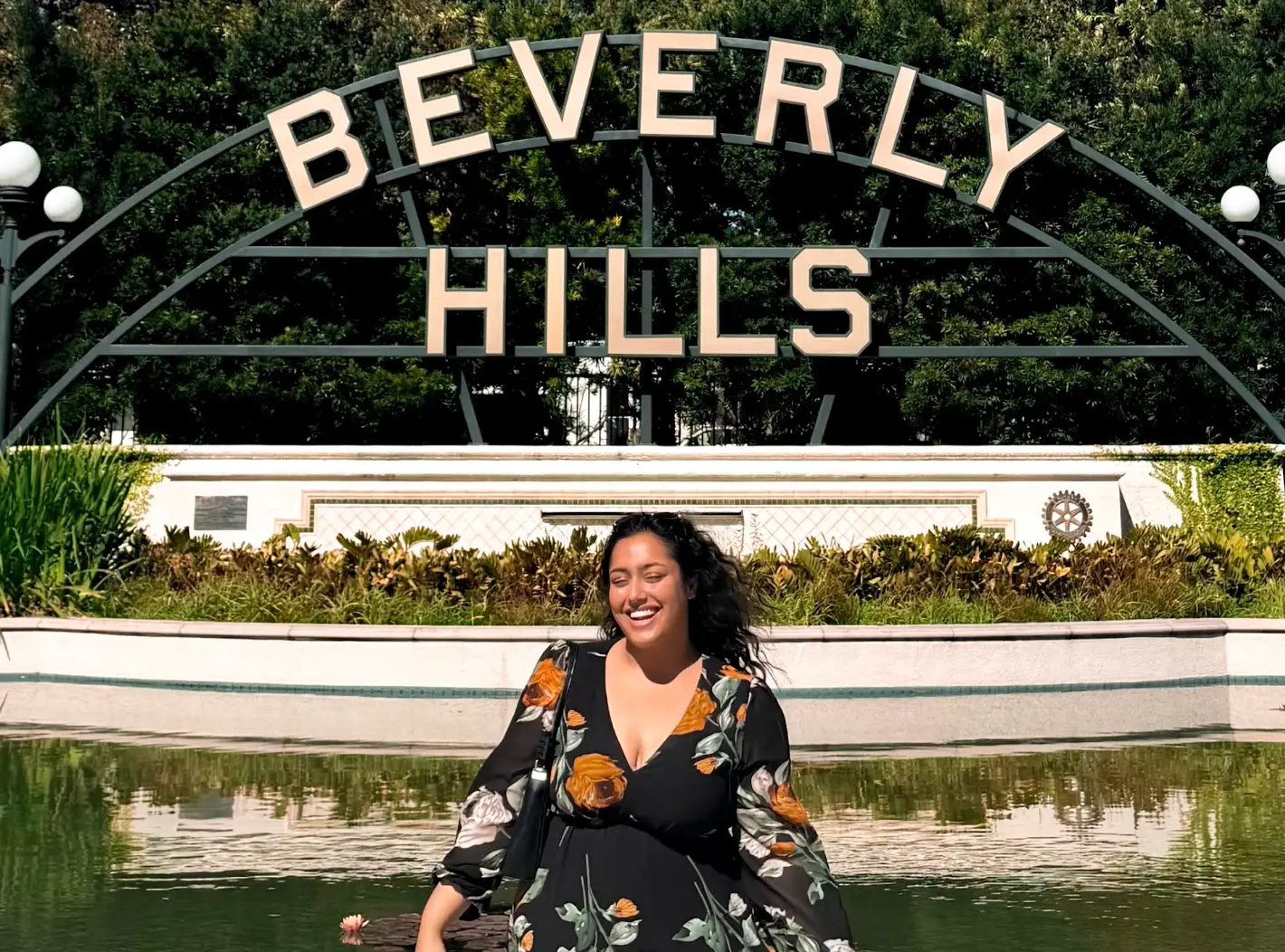 A smiling person stands in front of a Beverly Hills sign on a sunny day.