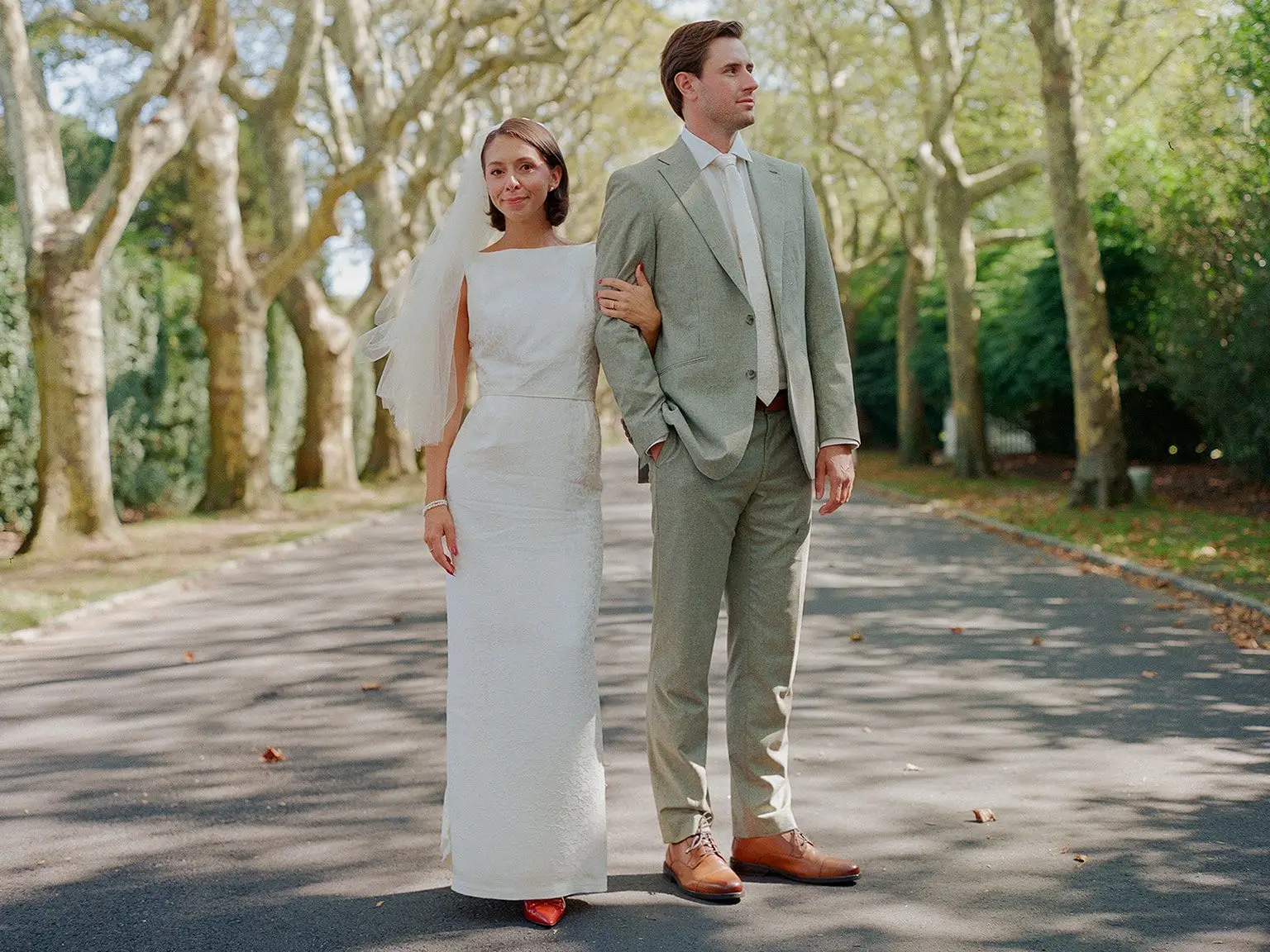 A bride and groom stand on a tree-lined street in their wedding attire.