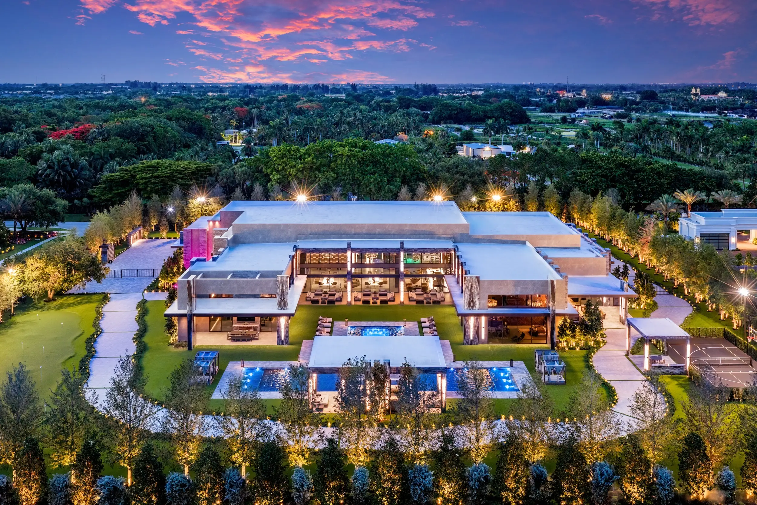 Aerial view of a large illuminated estate with pools, landscaped grounds, and surrounding trees at sunset.