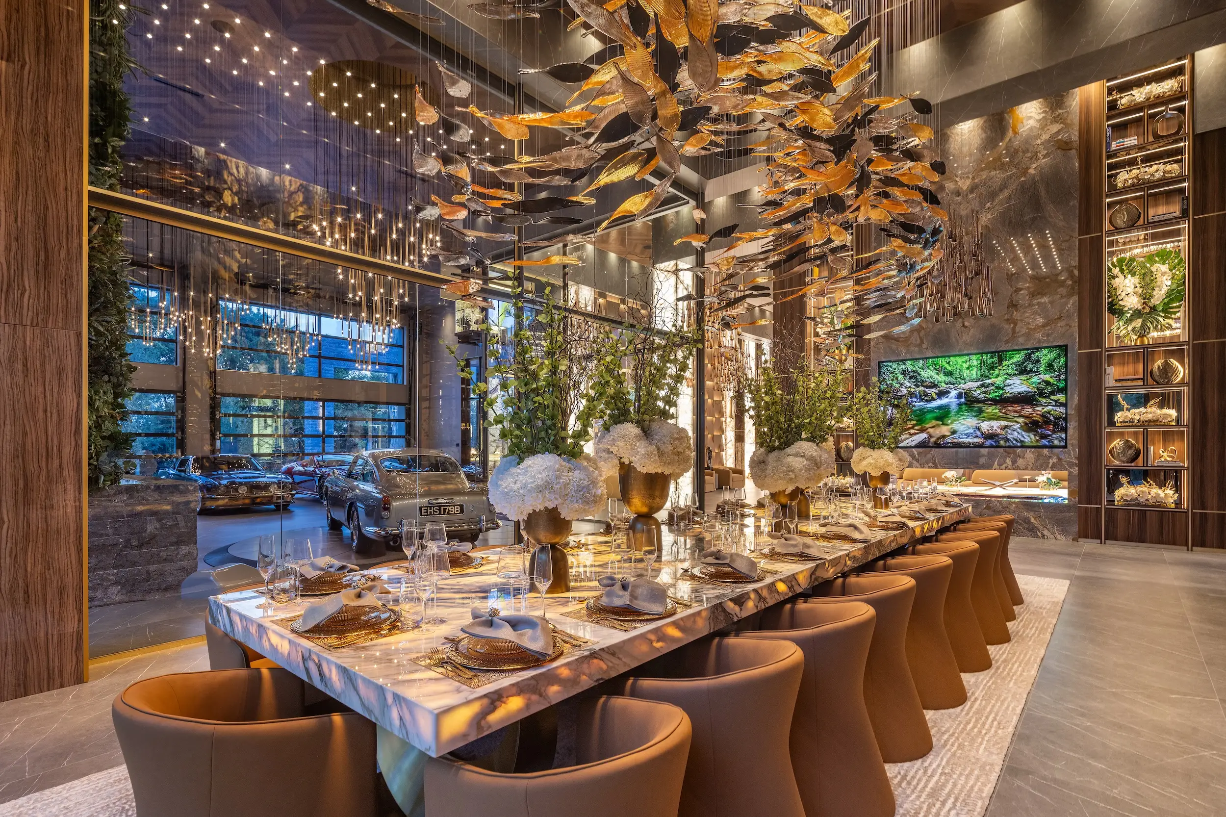 Luxurious dining room with a long set table, floral centerpieces, gold hanging decor, and cars visible behind glass.