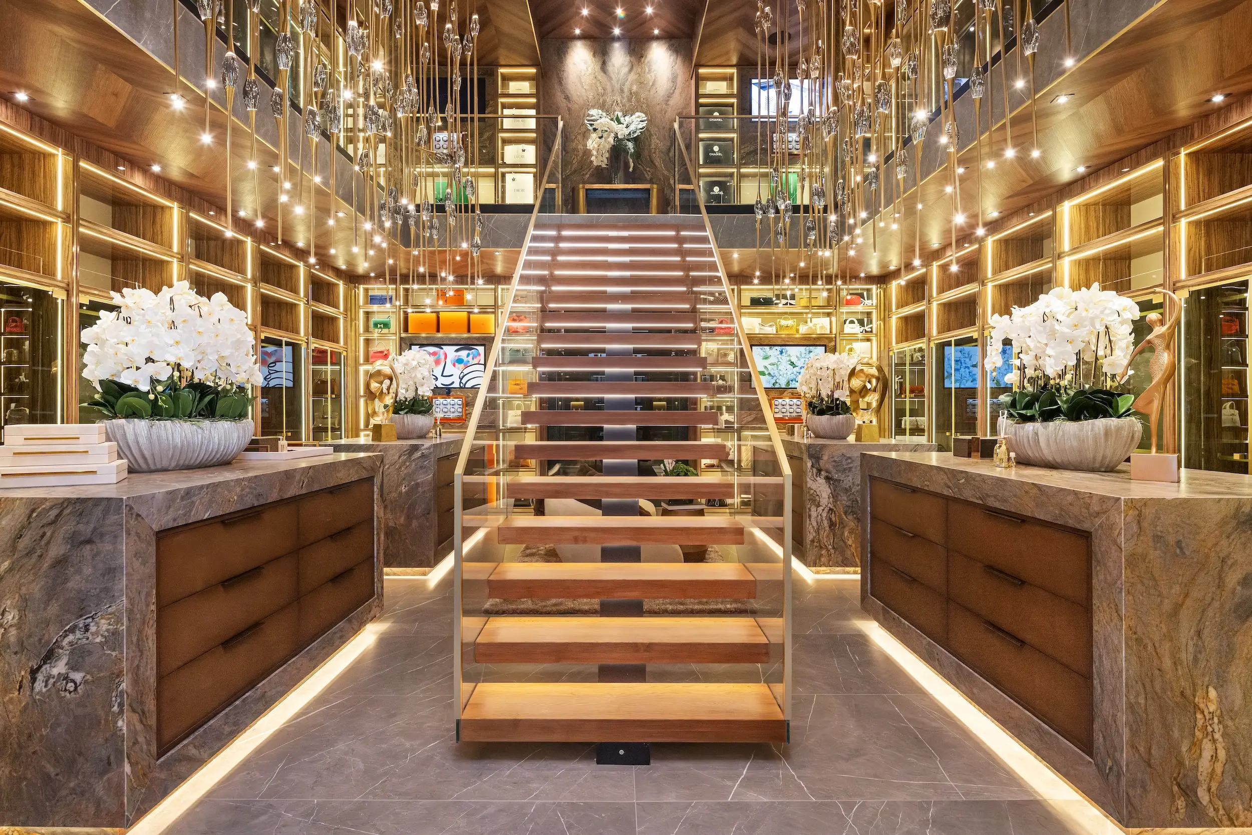 Luxurious boutique closet with illuminated central staircase, glass displays, marble counters, and white orchid arrangements.
