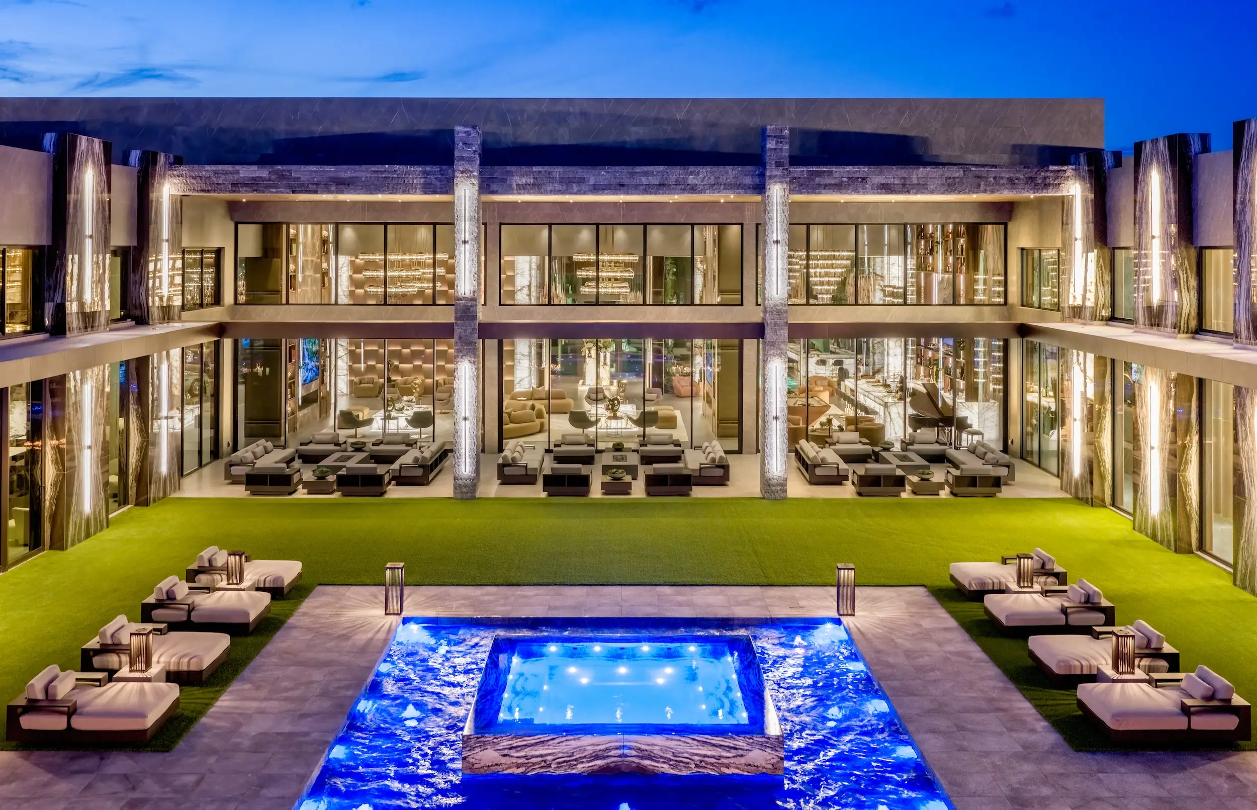 Illuminated modern luxury residence with glass walls, courtyard lawn, lounge beds, and a blue-lit swimming pool at dusk.