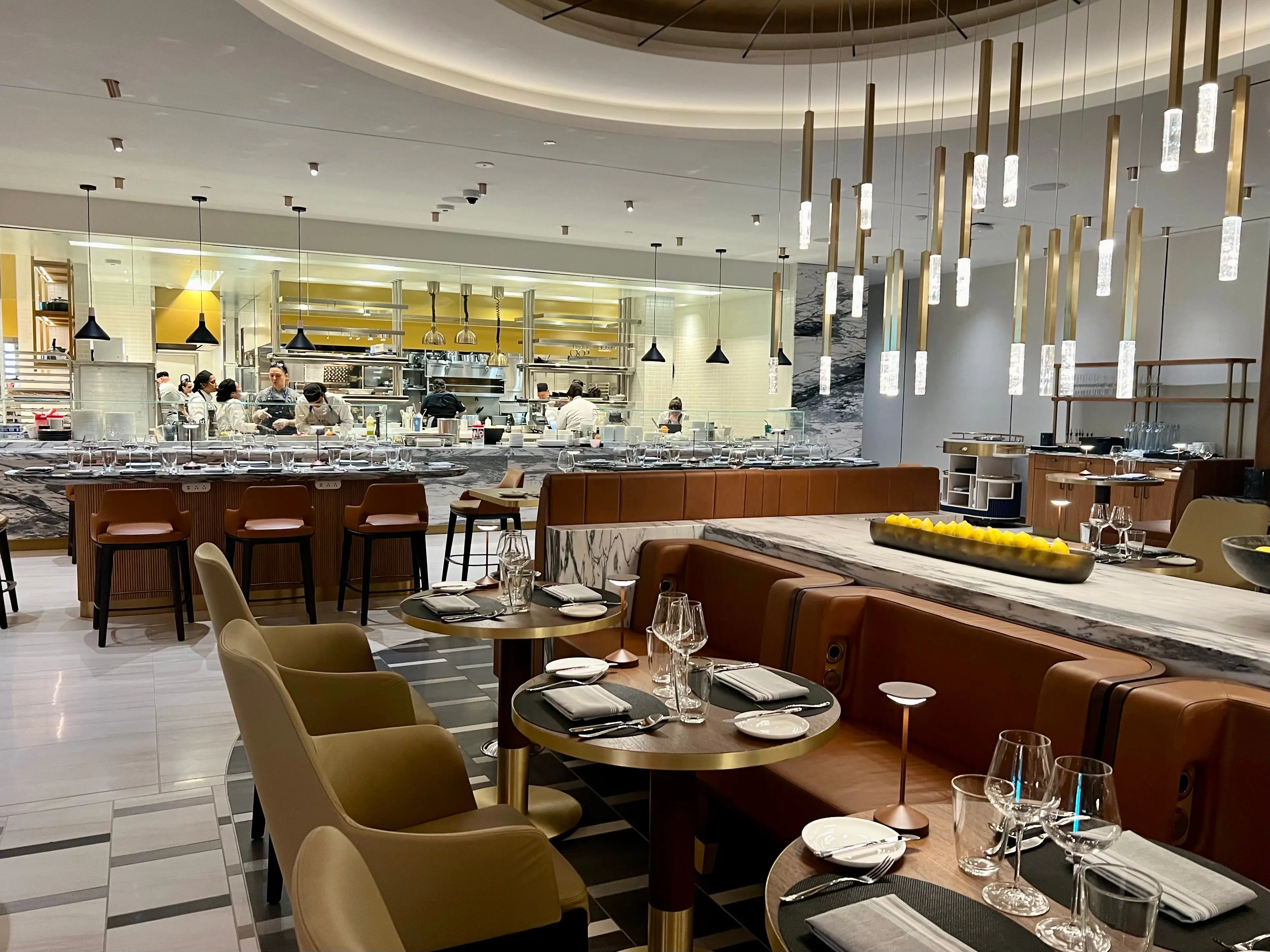 The Delta One Lounge Brasserie restaurant has beige and caramel-colored booths, chairs, and a hanging chandelier. The viewer is facing the open kitchen.