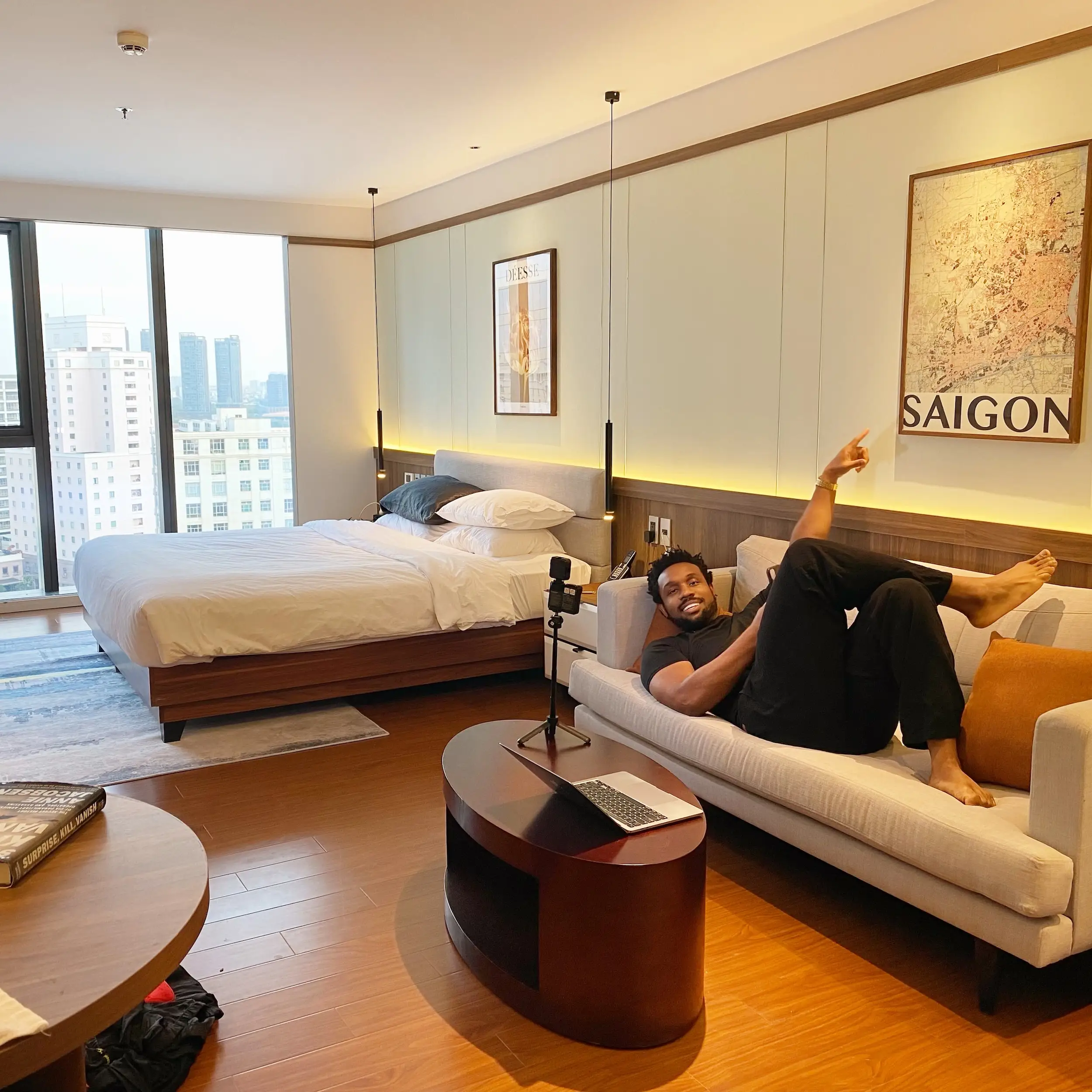 Person reclines on a sofa in a modern hotel room with a bed, city view, and SAIGON wall art.