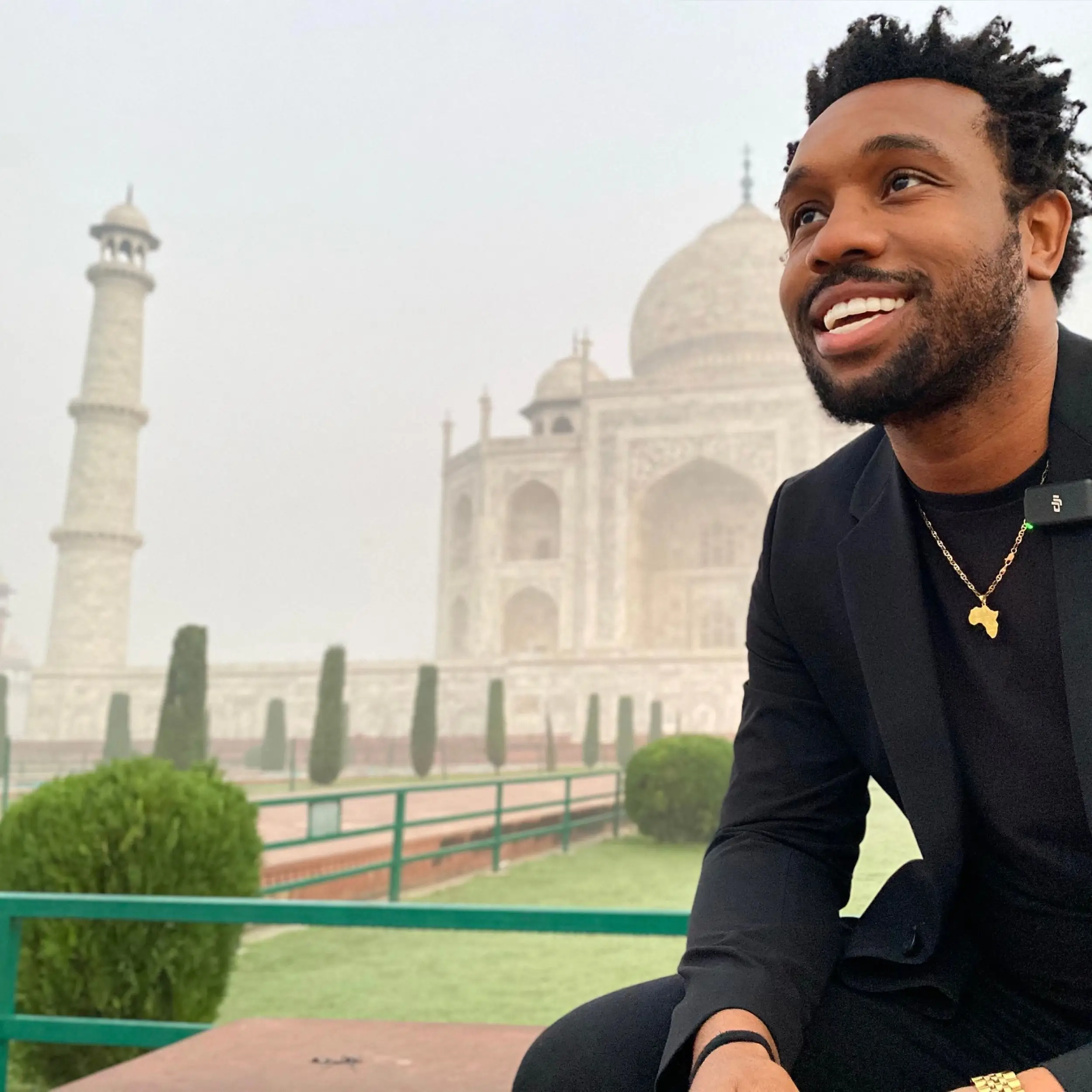 A smiling person in black clothing poses in front of the Taj Mahal on a misty day.