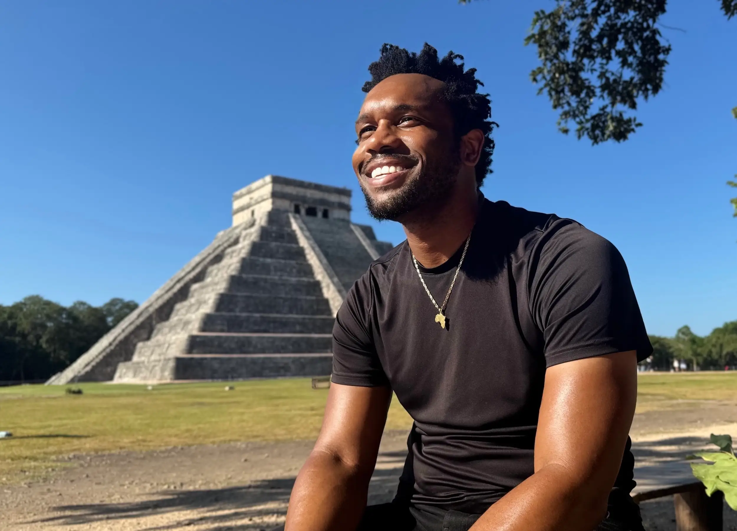 A smiling person seated outdoors in front of the El Castillo pyramid at Chichén Itzá under a clear blue sky.