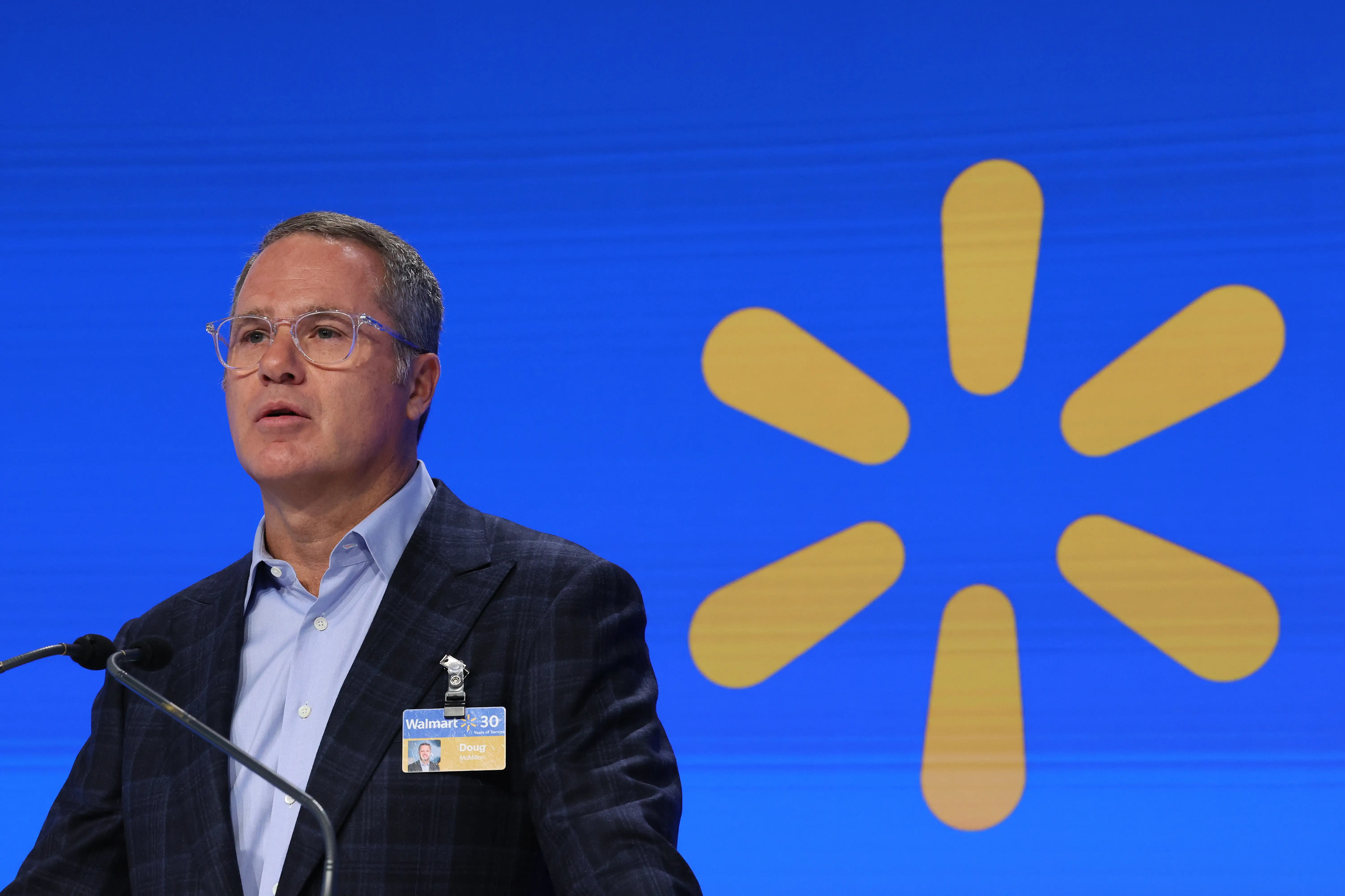 Former Walmart CEO Doug McMillon speaks into a microphone in front of the Walmart logo.