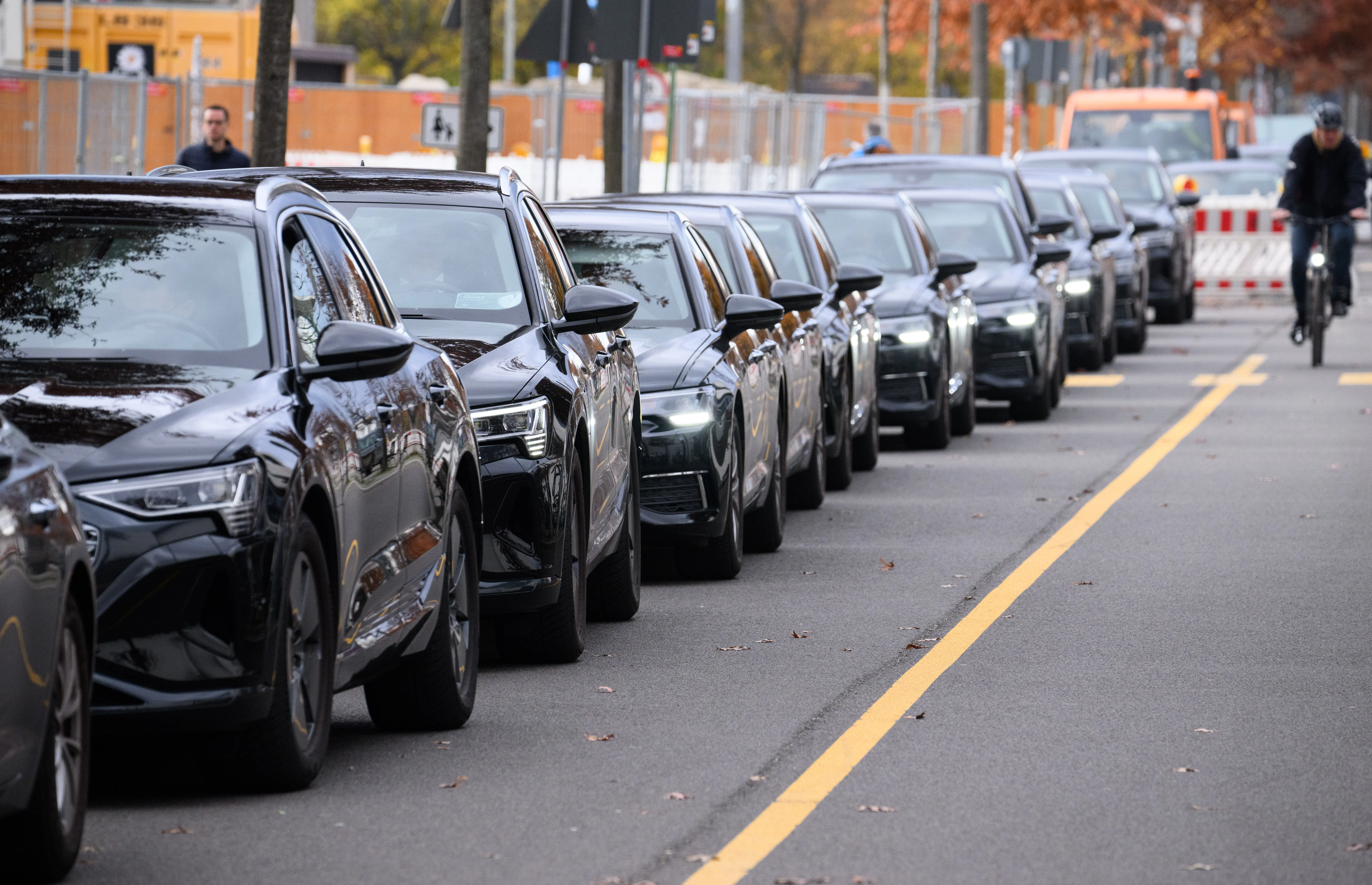 Black sedans and SUVs sit parked on a street as a cyclist rides by.