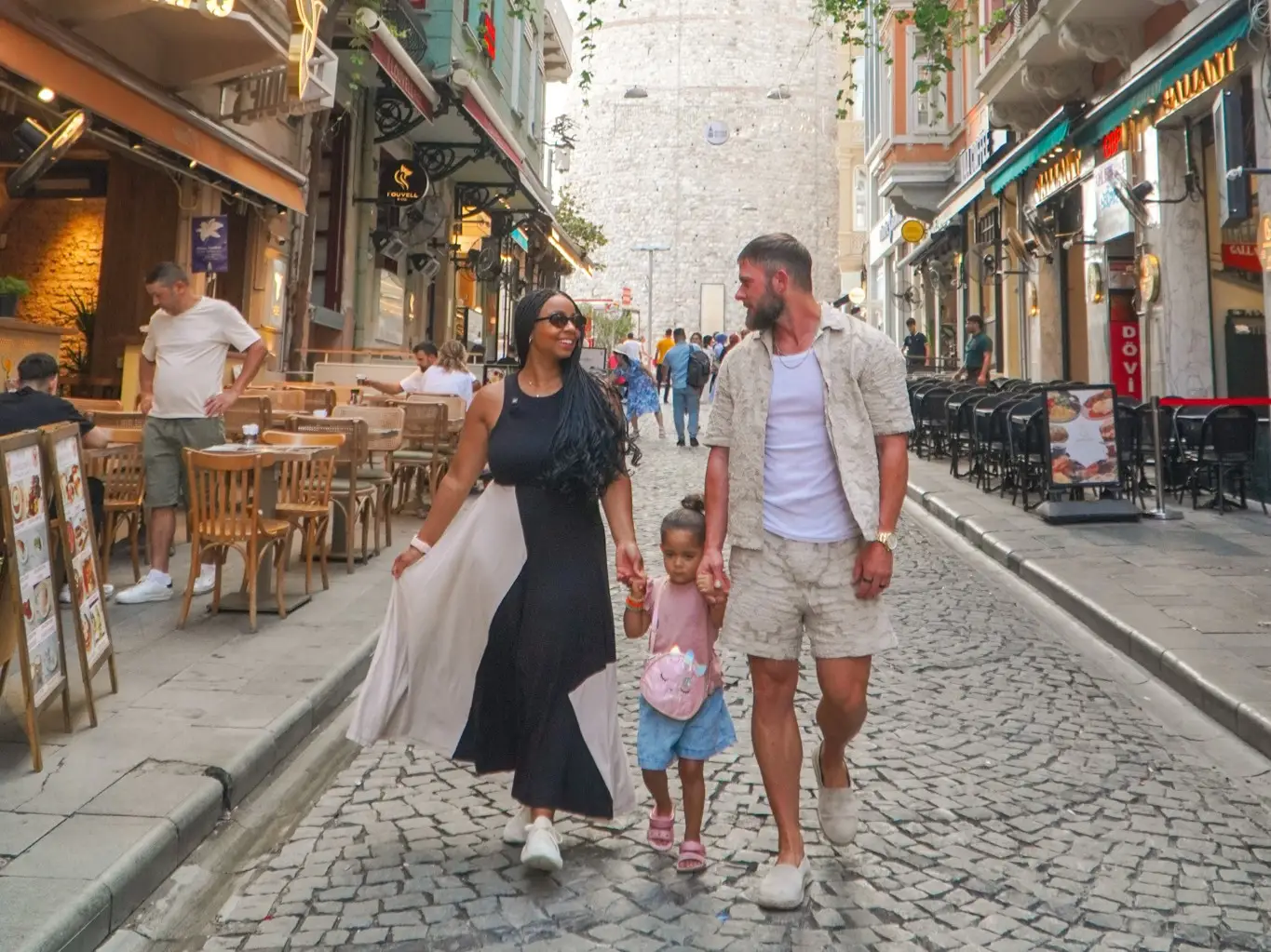 Three pedestrians walk along a cobblestone street with cafes beneath the Galata Tower in Istanbul.