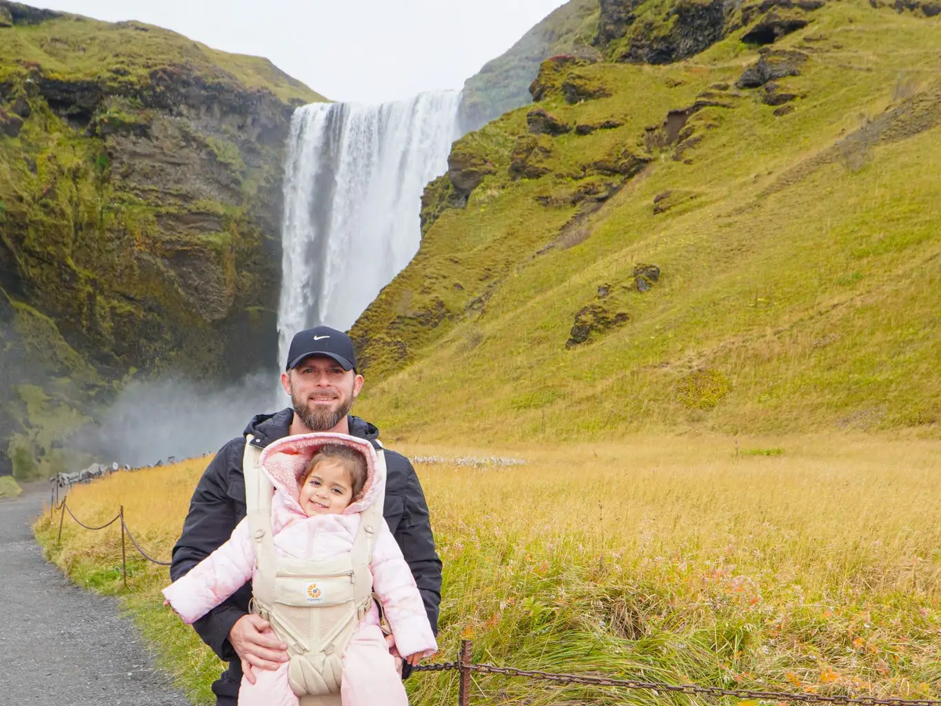 An adult carries a bundled child on a path below Skógafoss waterfall in a grassy canyon.