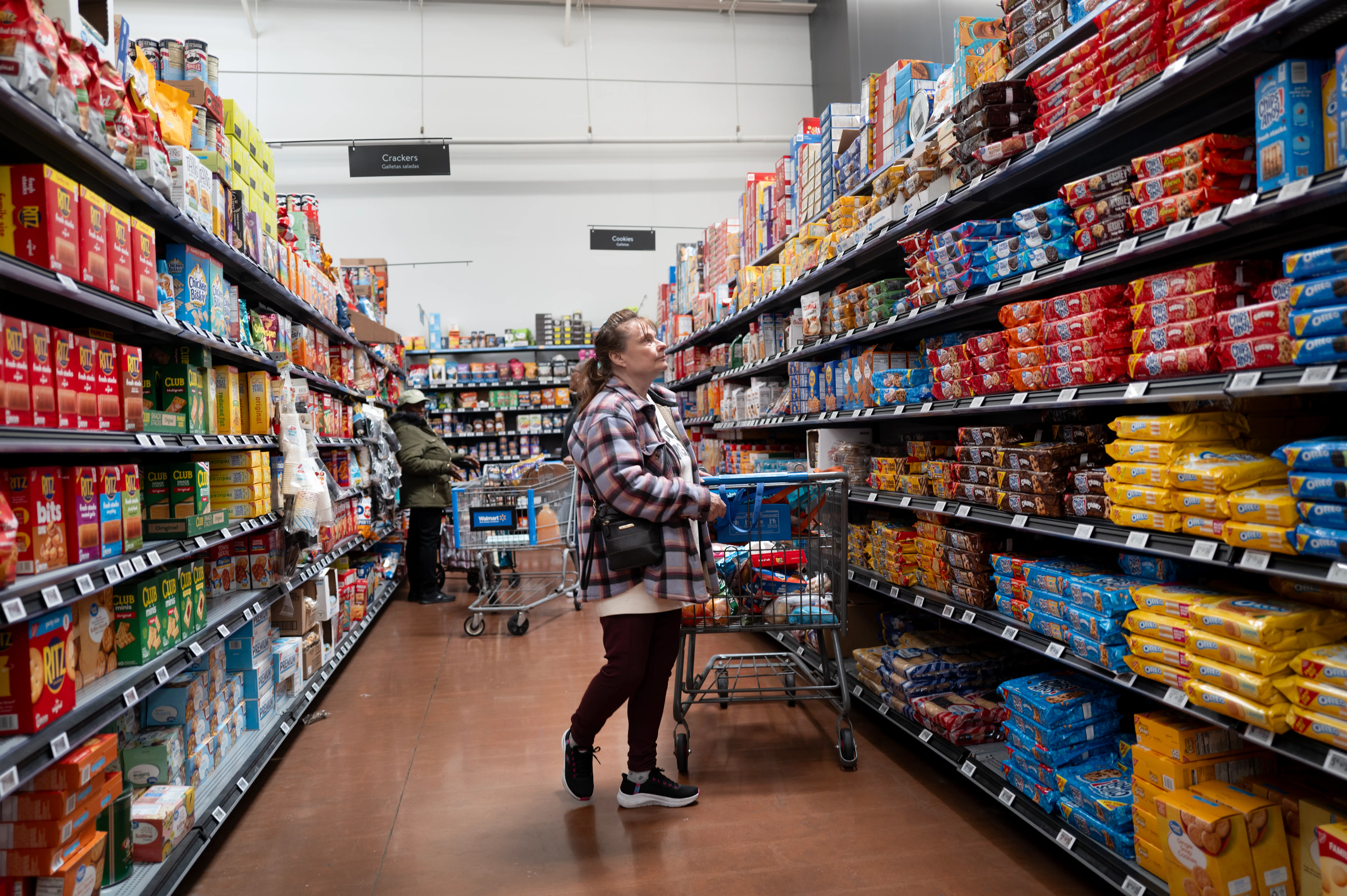 A woman stands with a shopping cart in the middle of an aisle where the shelves are stocked with packaged cookies at a Walmart store.