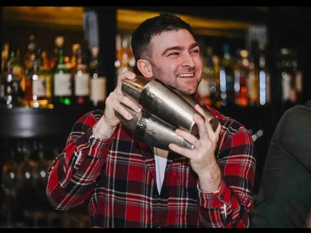 A bartender in a red flannel shirt is shaking a cocktail.