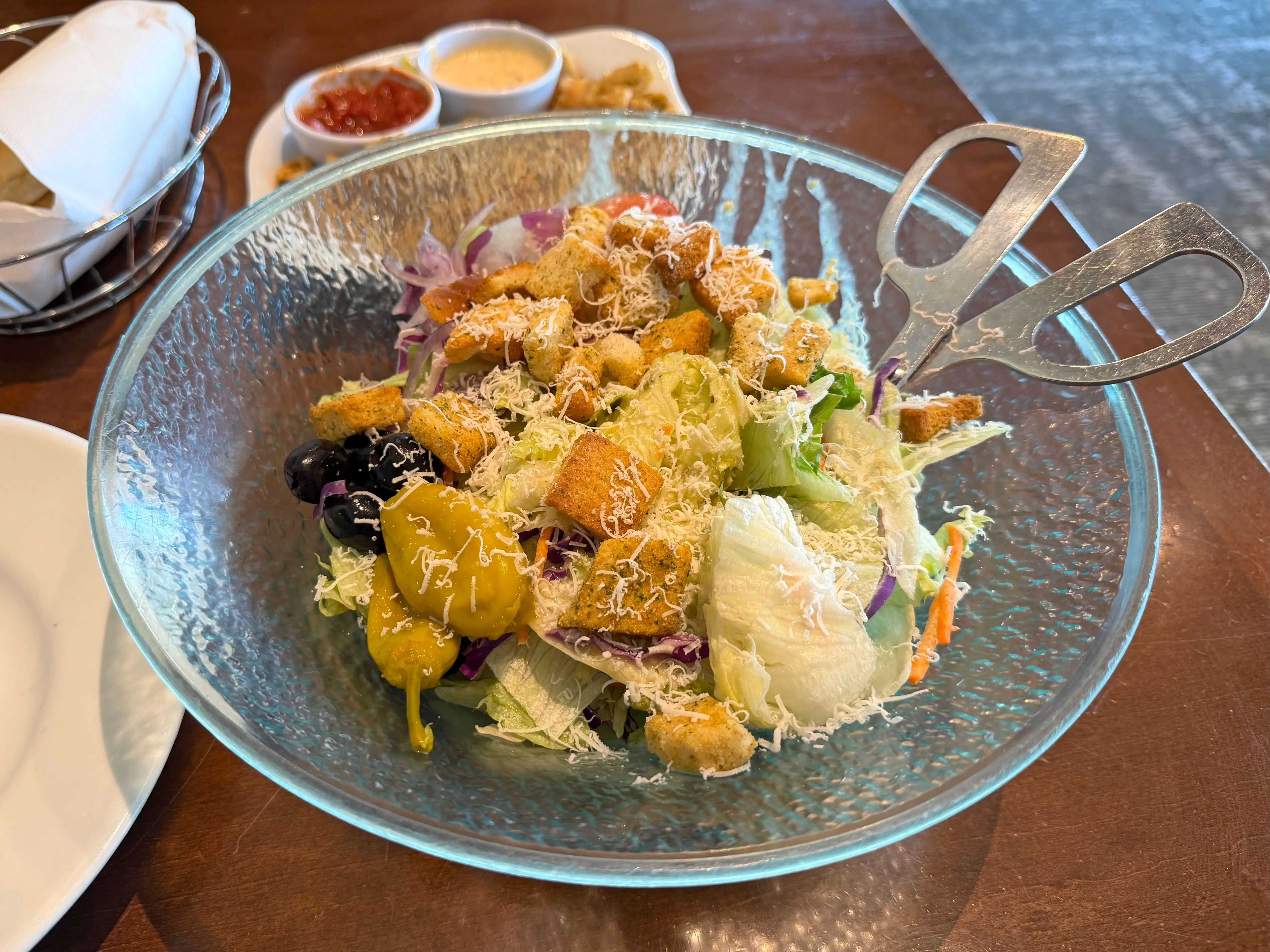 A large bowl of salad with lettuce, purple cabbage, shredded carrots, peppers, olives, croutons, and cheese.