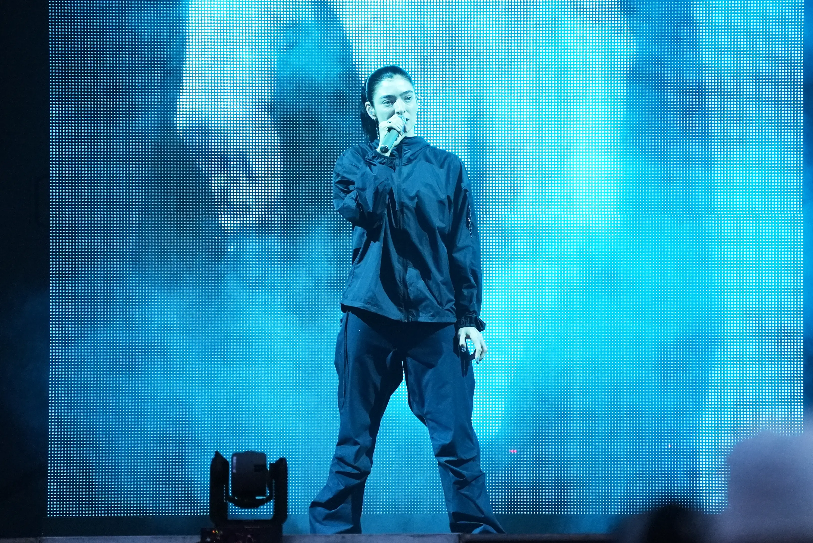 Lorde is holding a microphone in her right hand onstage.