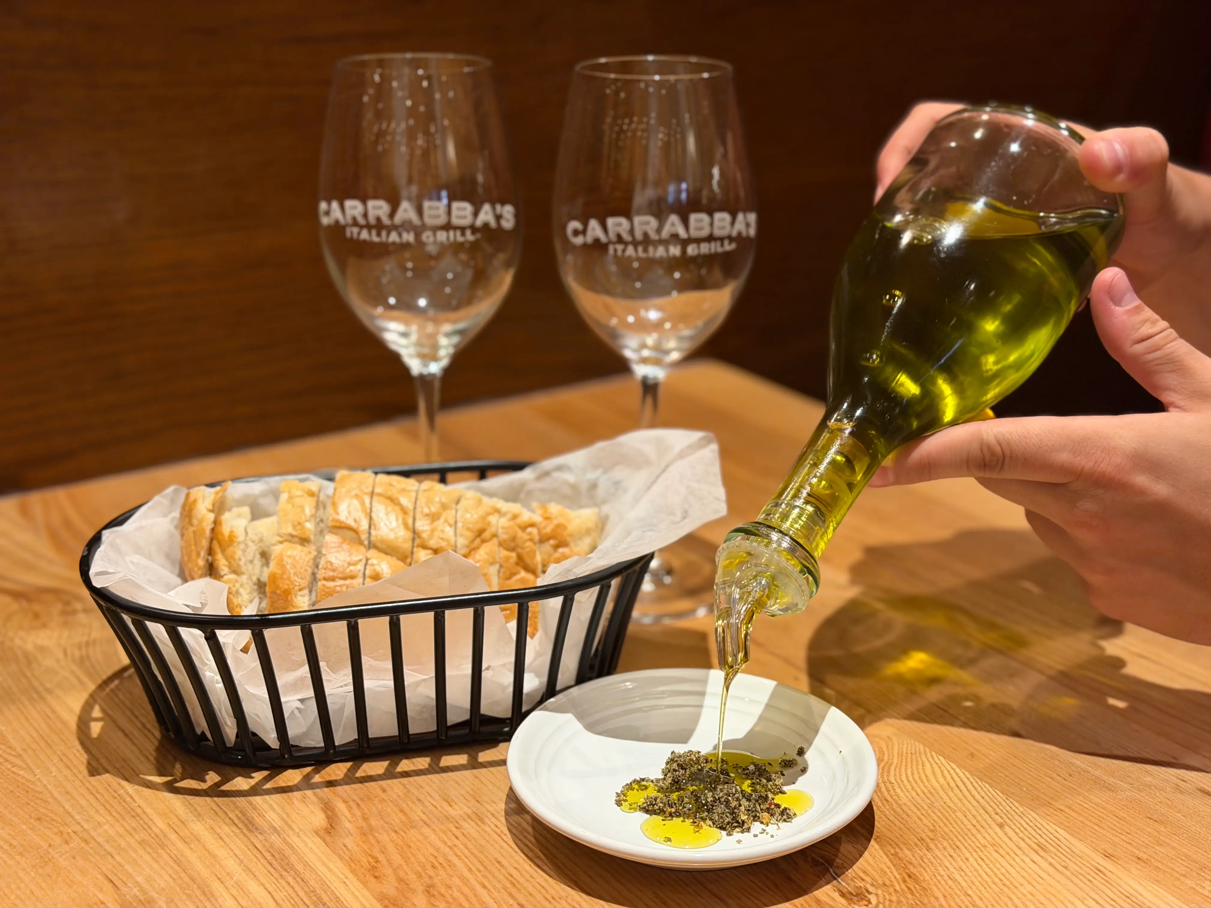 A person pours olive oil onto a white plate with herbs. There is also a basket of bread and two wine glasses that read