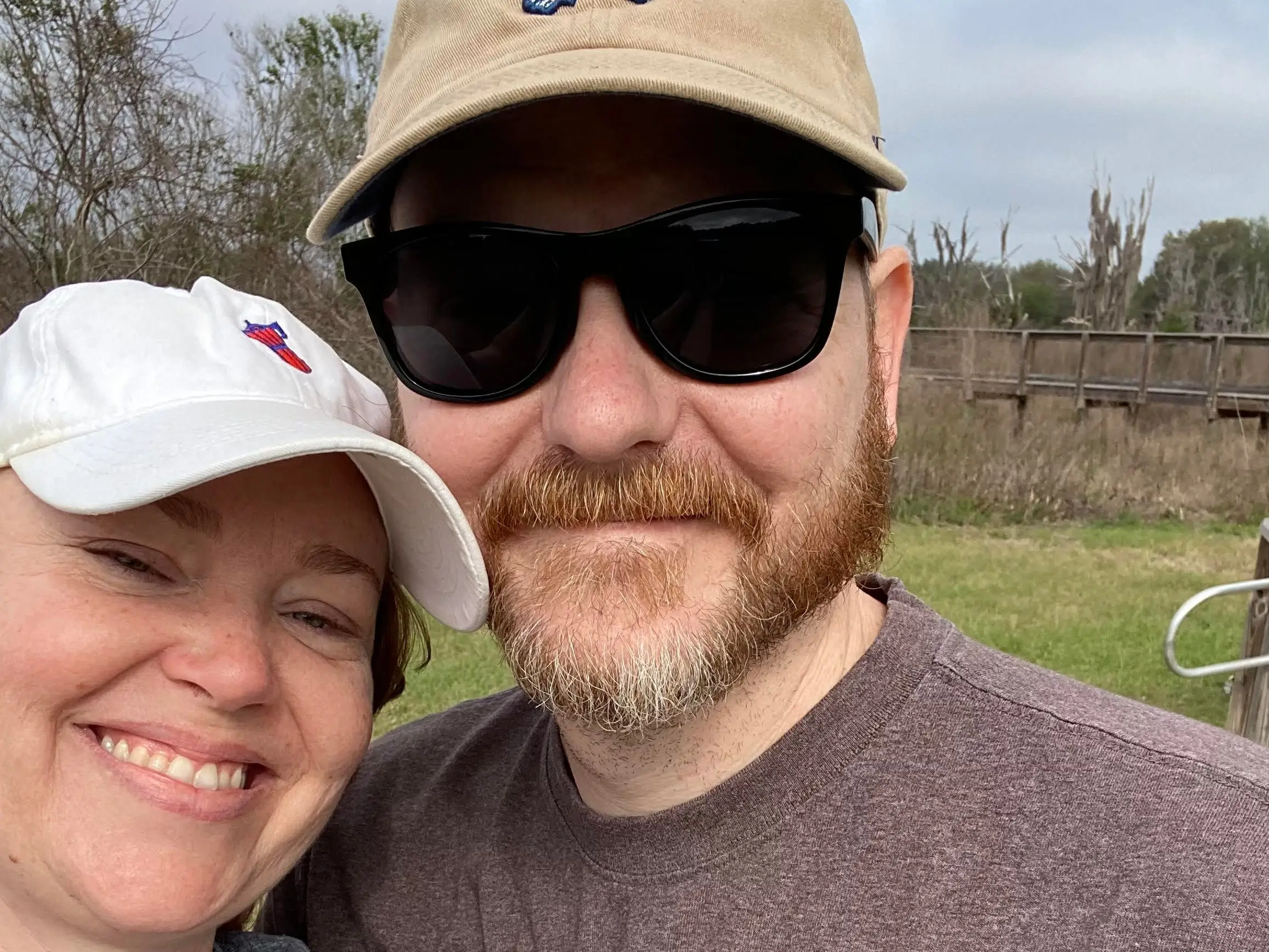 Two people wearing caps and T-shirts smile for a close-up outdoor selfie near grass and a wooden boardwalk.