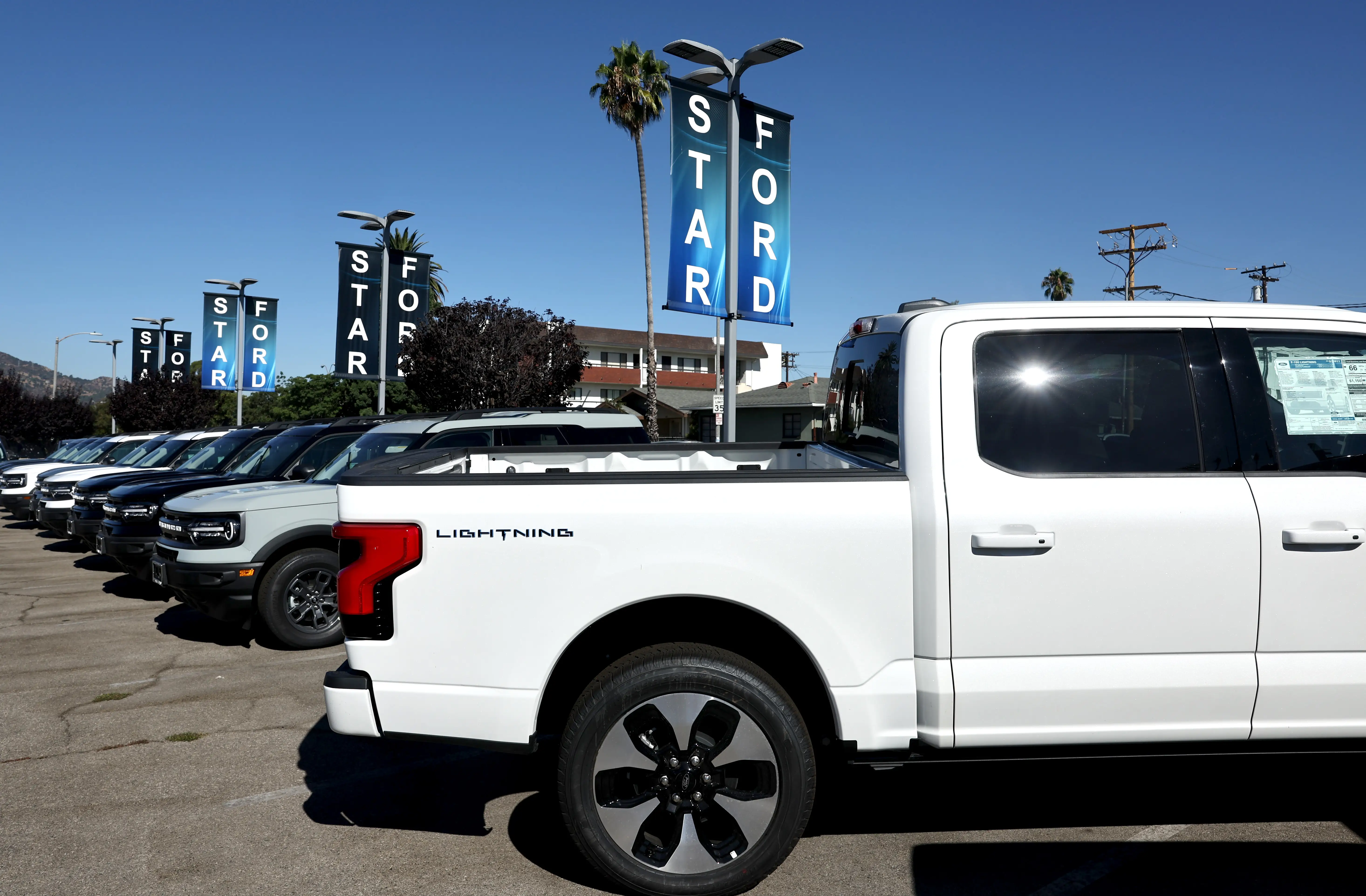 A white Ford F-150 Lightning on a dealership lot next to several gas-powered Ford Bronco Sport SUVs.