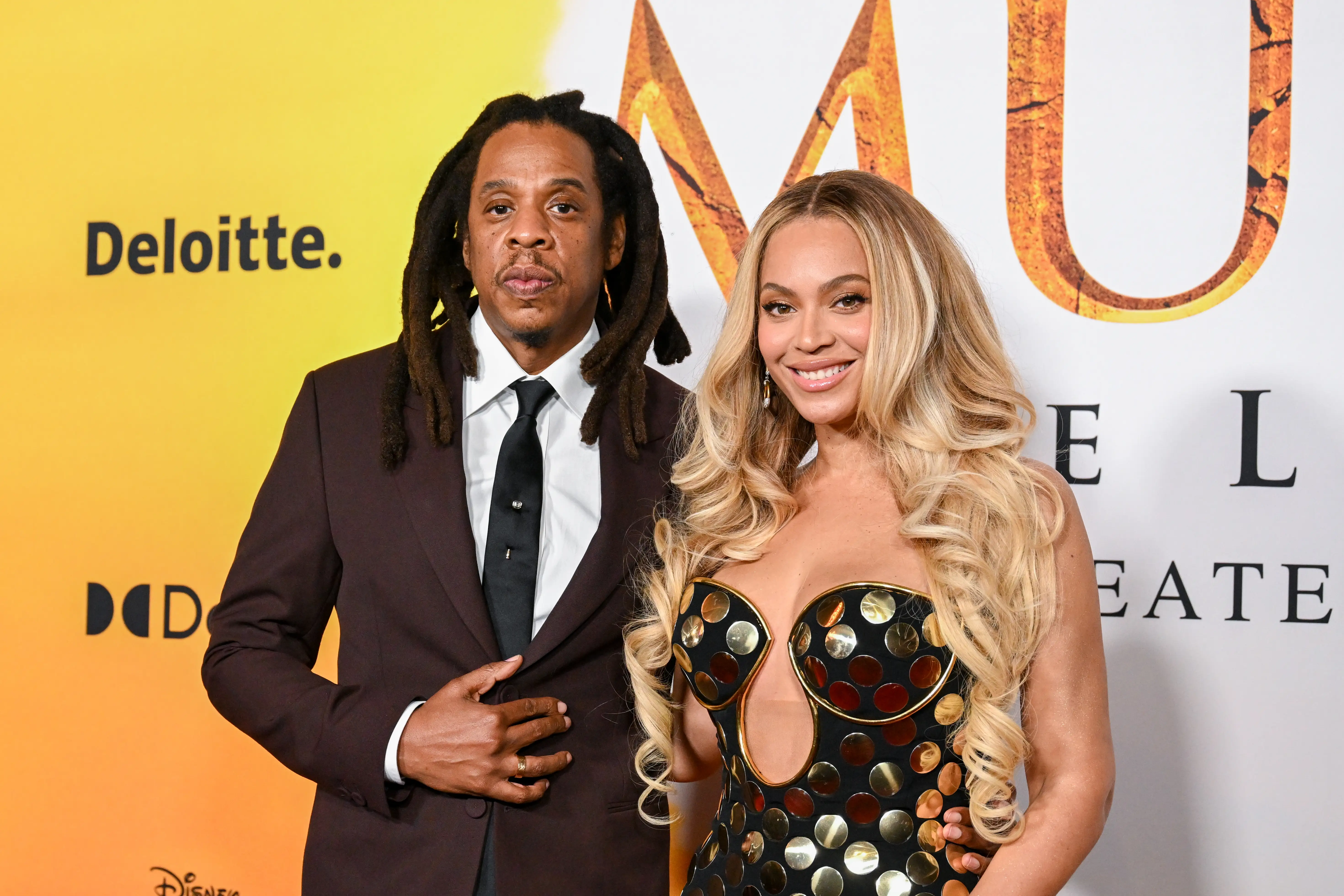 Jay-Z and Beyoncé pose together in formal attire before a branded event backdrop with Deloitte and Disney logos.