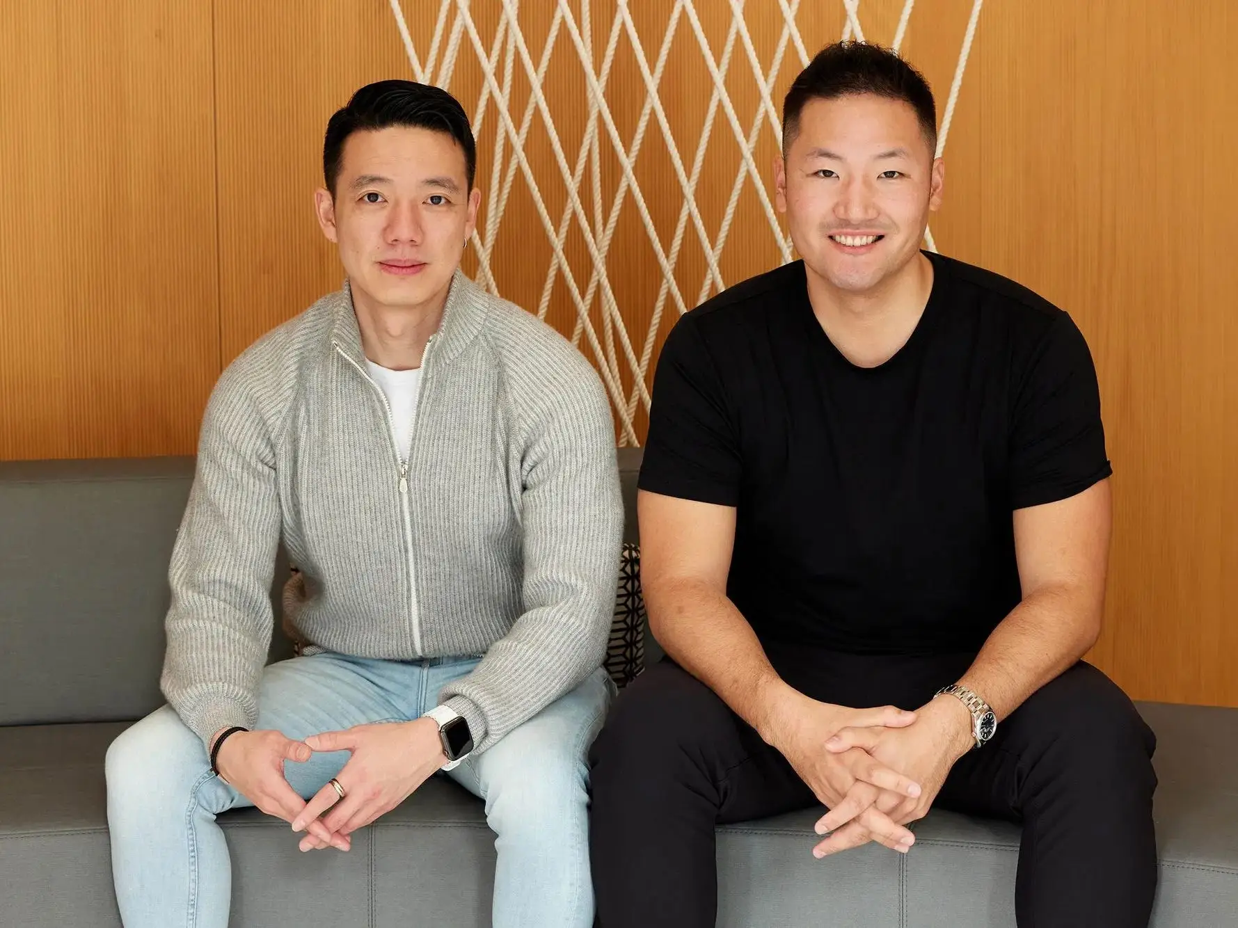 Two men sit on a gray couch in front of a wood-paneled wall with a white geometric rope pattern.