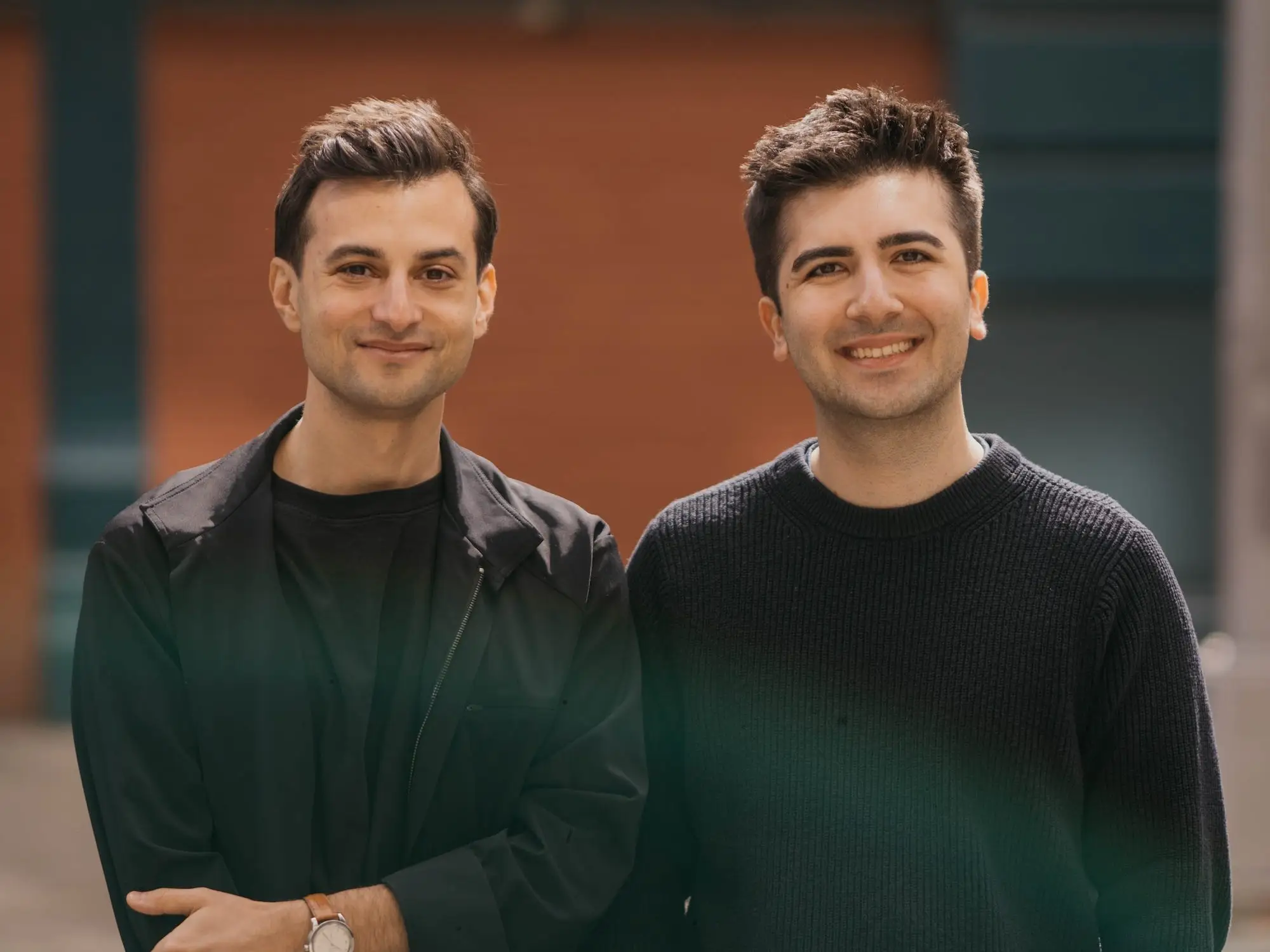 Two men in dark clothing stand outdoors on a city street with a softly blurred building behind them.