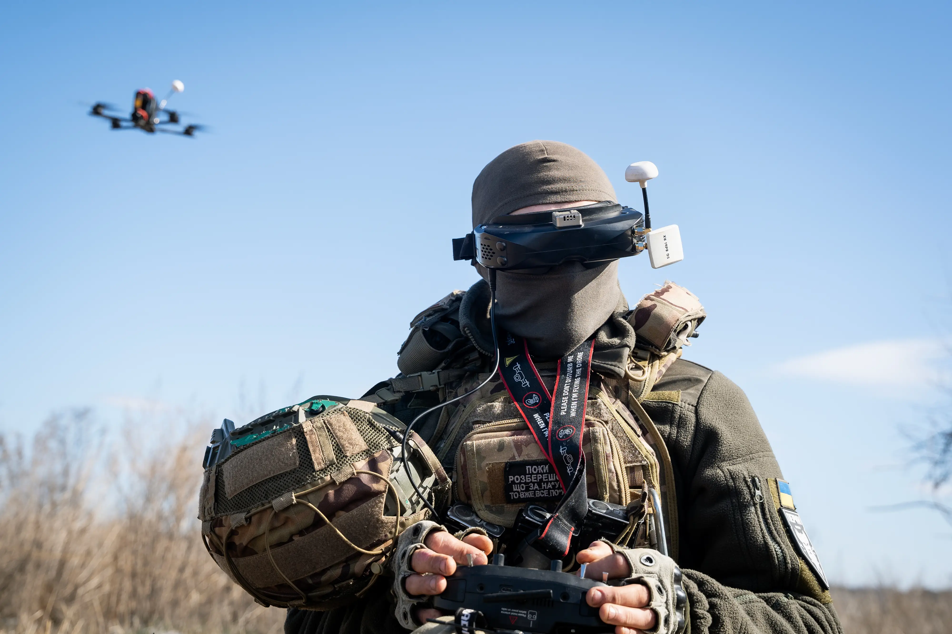 A man wearing khaki gear including a face covering wears eye goggles and holds a controller with a drone flying in a blue sky behind him