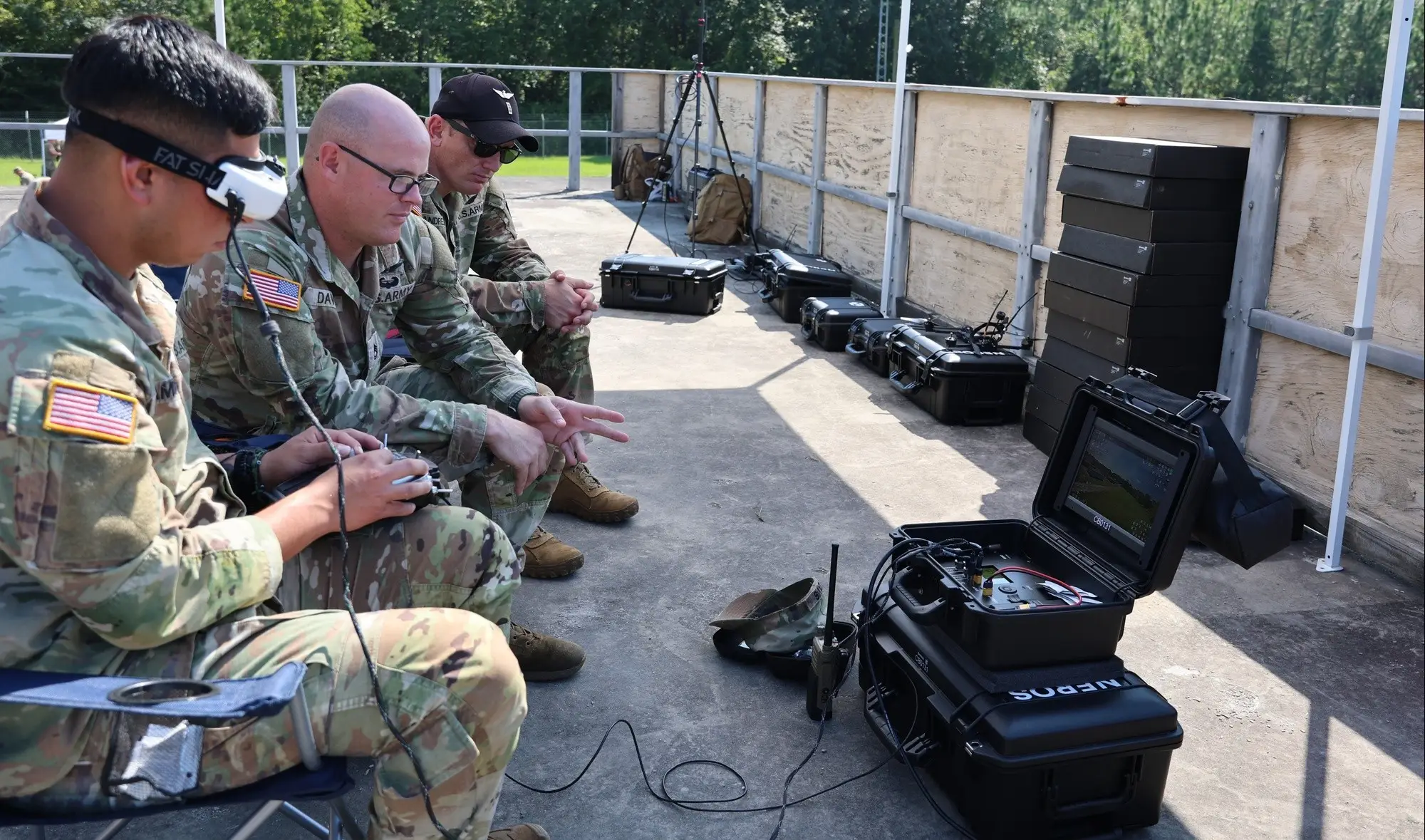 Military personnel in camouflage uniforms sit beside portable electronic equipment cases during an outdoor training setup.