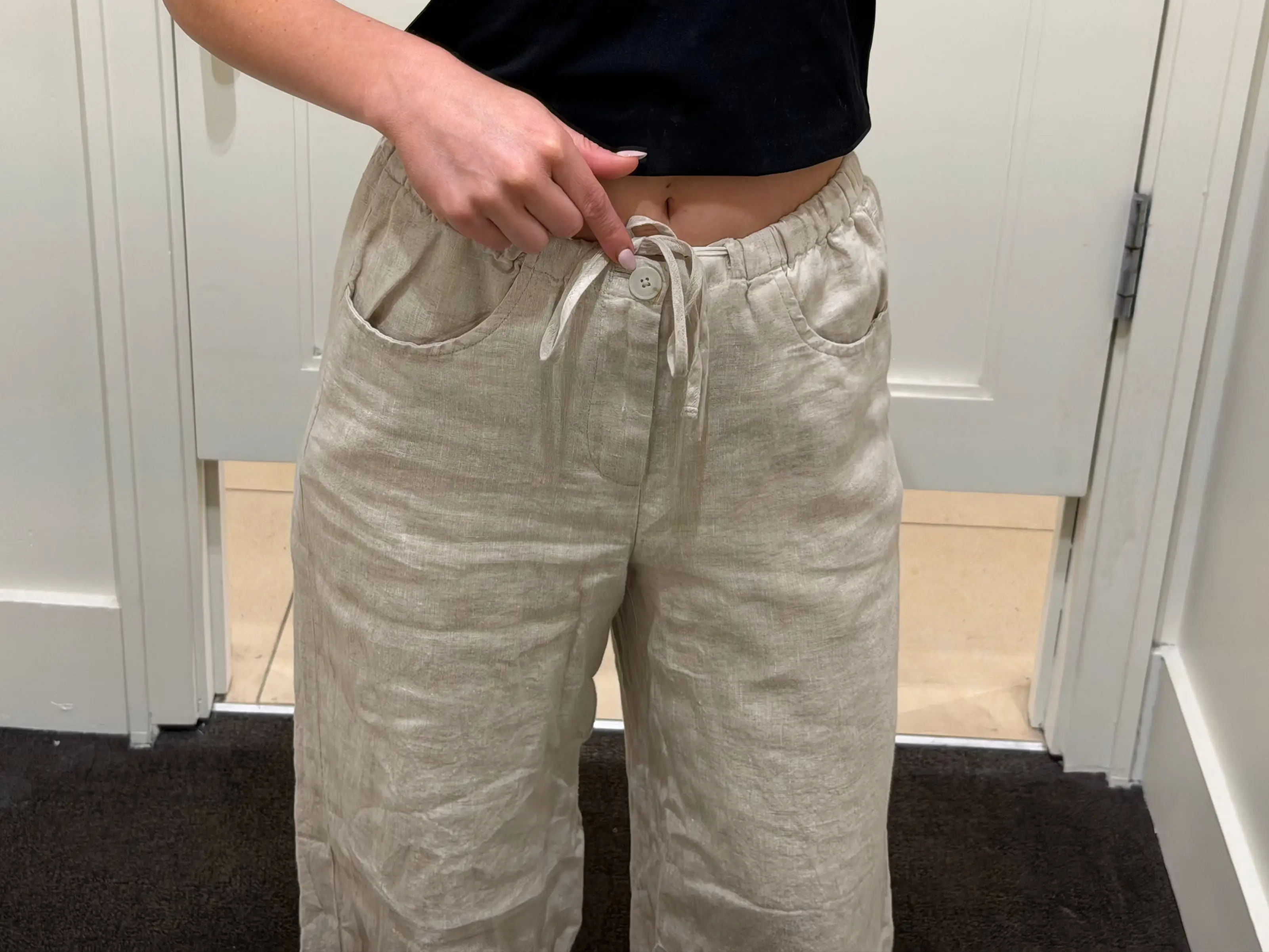 Chloe wears a pair of beige linen pants and points to the button.