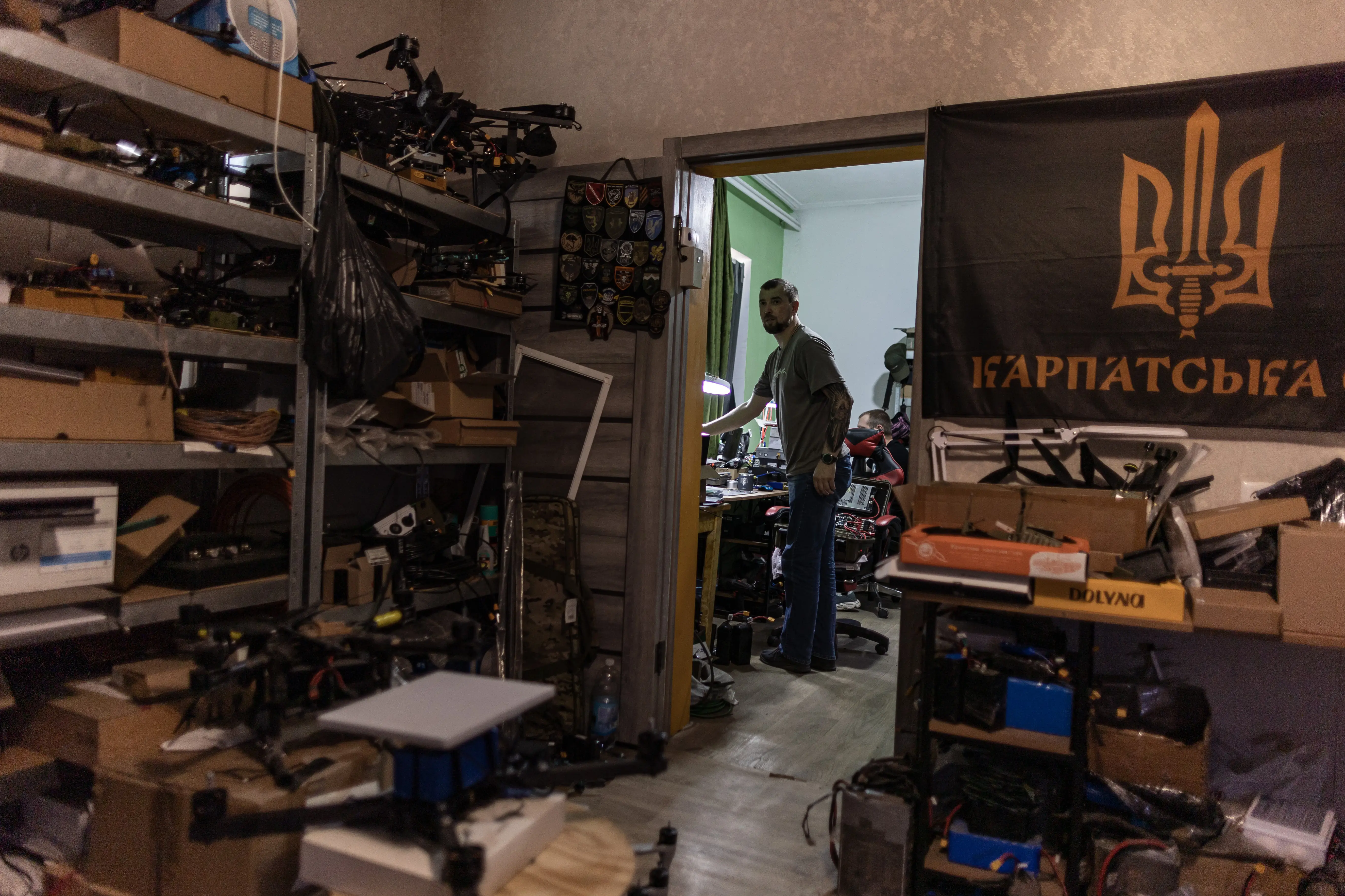 A room with a large flag and shelves of drone parts, with a doorway and a man standing in it visible