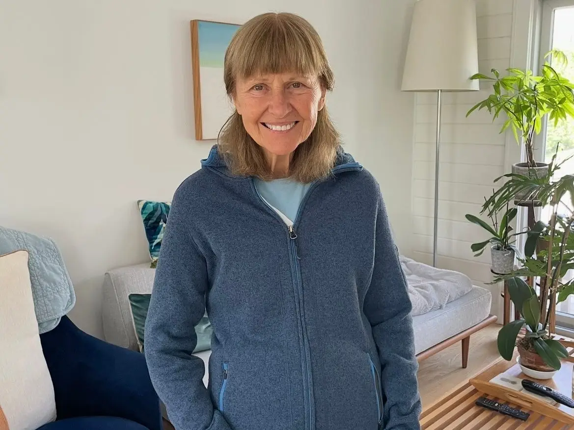 Smiling person in a blue zip-up fleece stands in a bright living room with plants and seating.