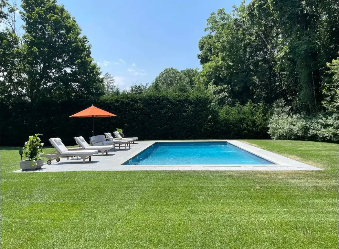 Backyard swimming pool with lounge chairs and an orange umbrella surrounded by lawn, hedges, and tall trees.