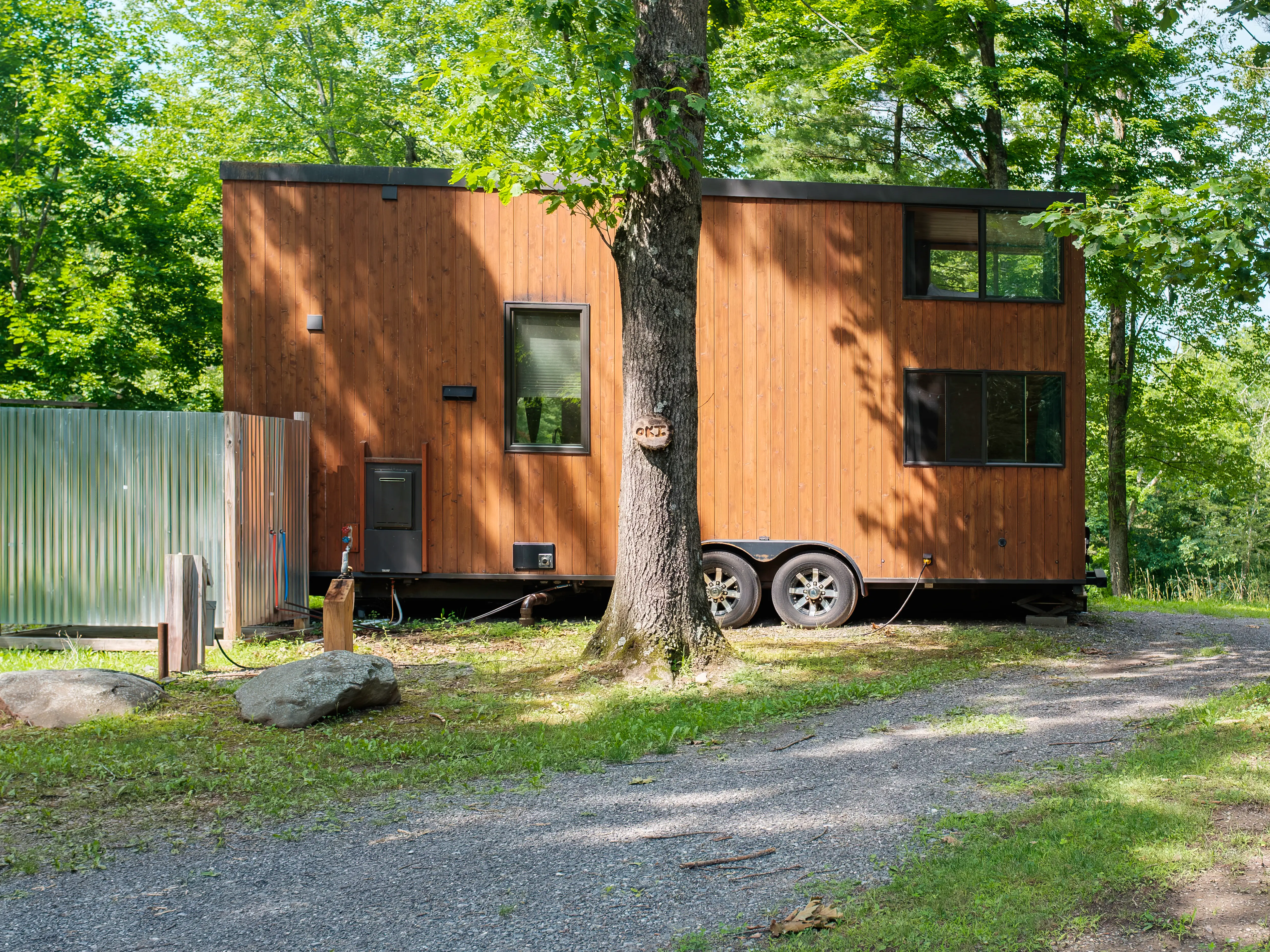 A wood-sided tiny house on a trailer sits beside a gravel driveway in a leafy wooded setting.